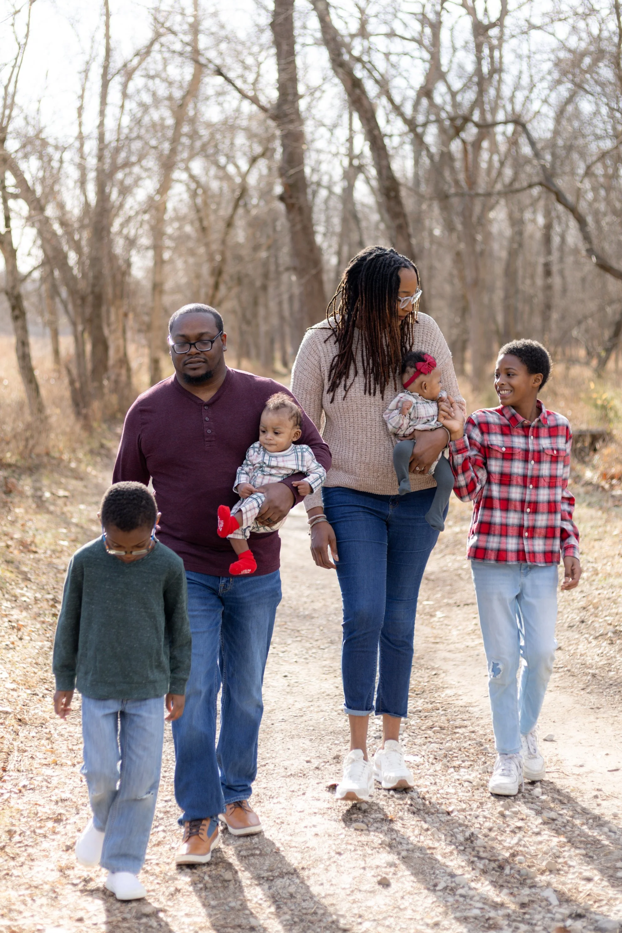 A family of six walking on a dirt path through a wooded area during autumn. The father is carrying a toddler girl, while the mother is holding a baby girl. A teenage boy and a young boy walk beside them, all engaged in conversation and enjoying the outdoors.