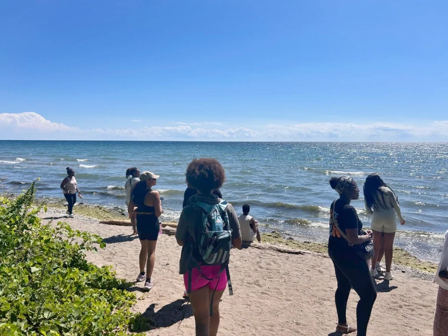 People on a sandy beach near the ocean under a clear blue sky, some standing and some walking by the water.