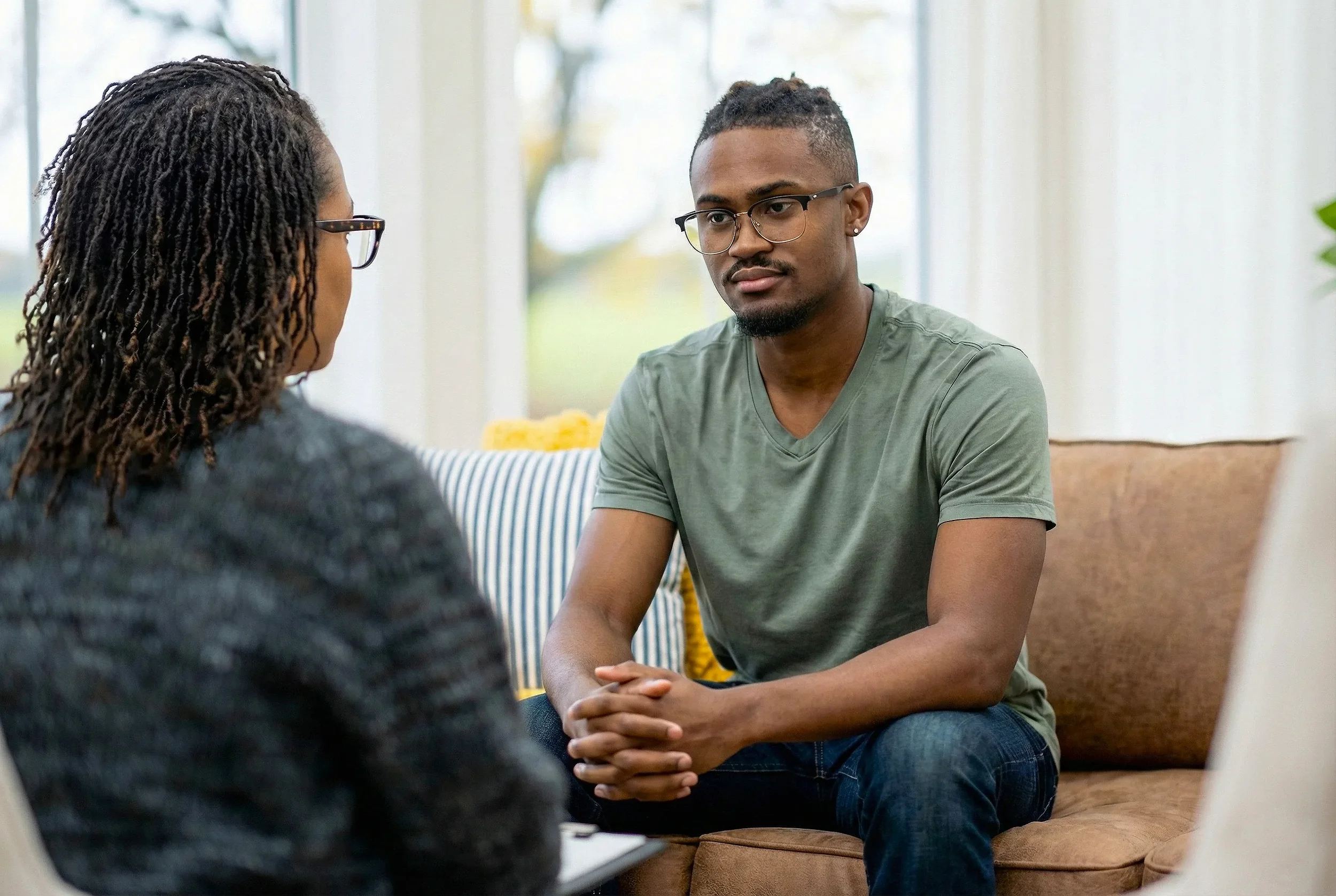 A young man with glasses and a green t-shirt sitting on a couch during a conversation with a woman with glasses and braids, indoors with large windows in the background.