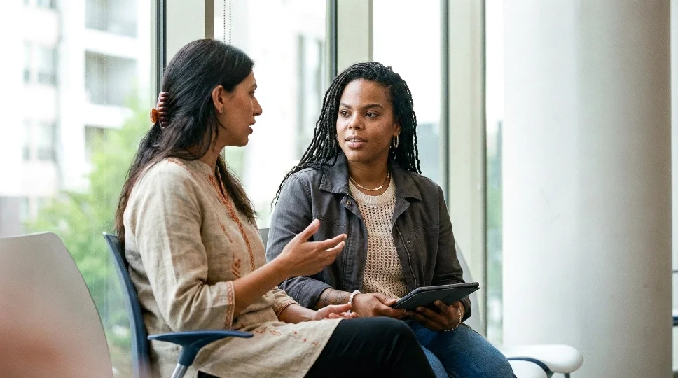 Two women sitting and talking in a waiting room near large windows