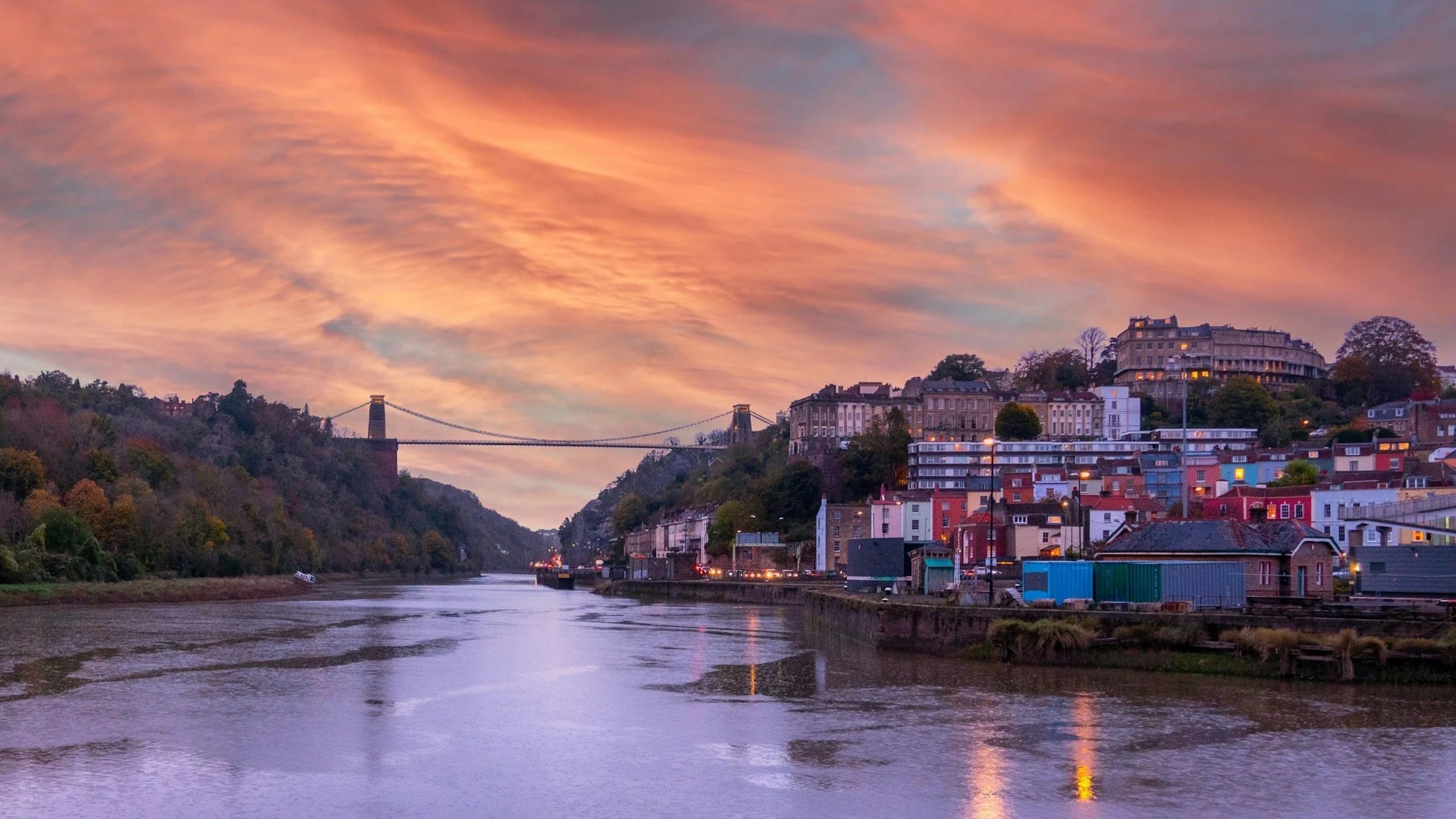 Sunset over a river with colorful clouds, a bridge connecting two hills, and a hillside town with houses and buildings.