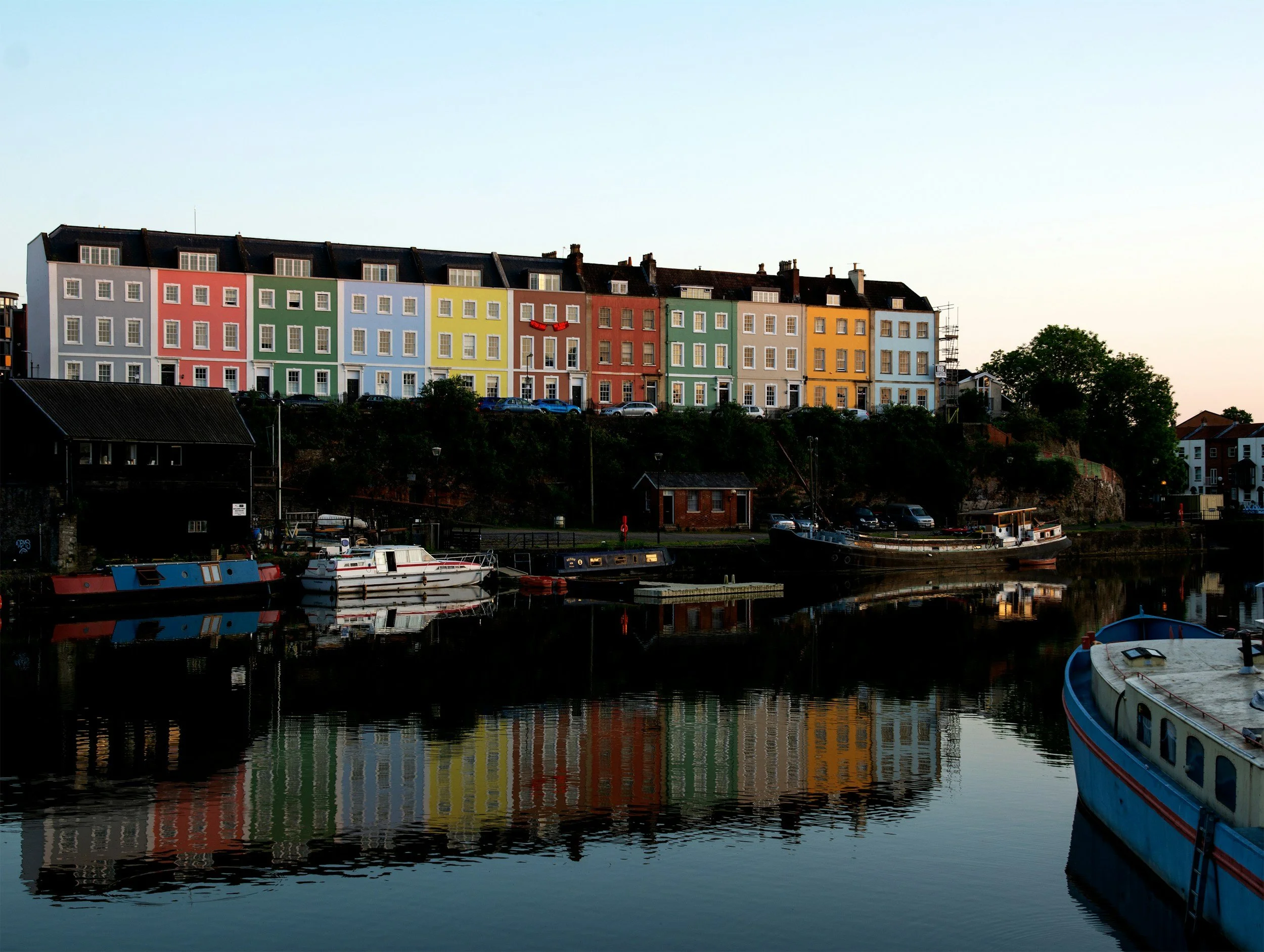 Colorful apartment buildings overlooking a river with boats docked along the water at sunset or early evening.