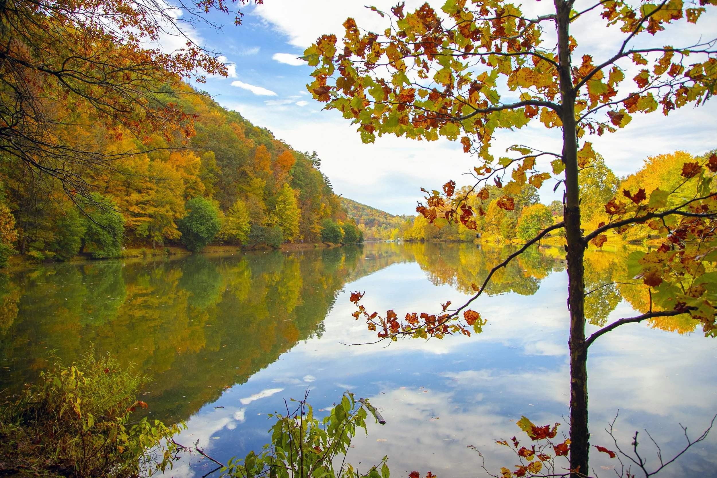 Scenic view of a calm river reflecting a colorful autumn forest and a partly cloudy sky.