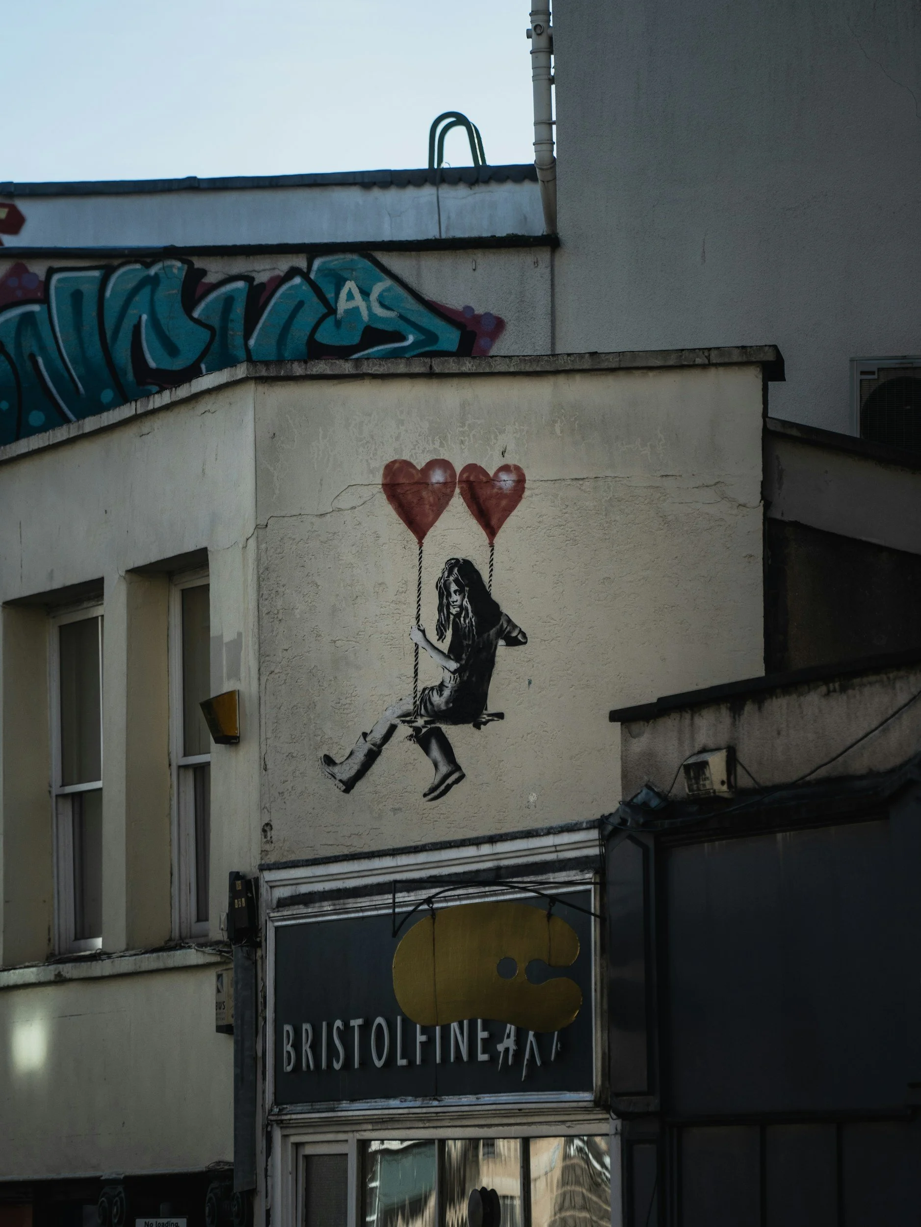 Street art mural of a girl sitting on a swing holding two red heart-shaped balloons, painted on a beige building wall. There is another building with graffiti and a sign for Bristol Fine Arts below the mural.