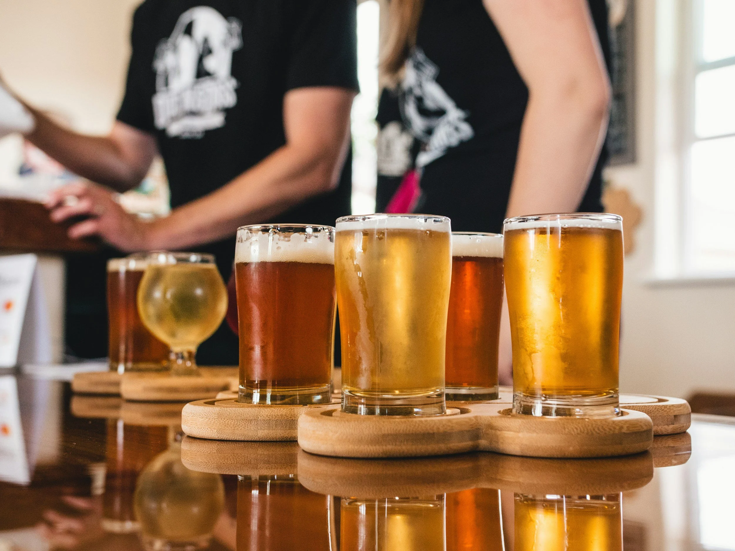 Various glasses of beer on a wooden table with people in the background.
