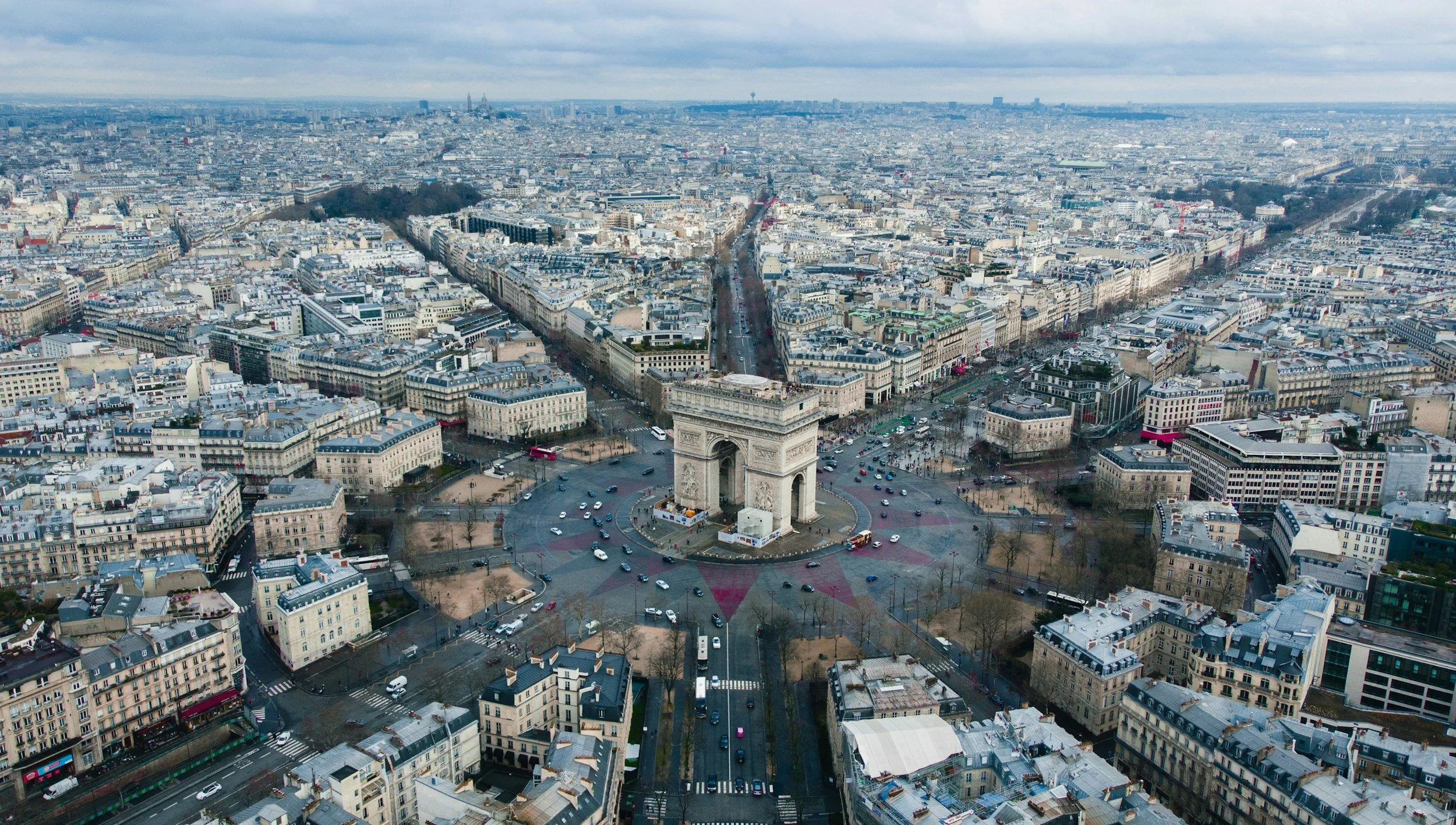 Aerial view of the Arc de Triomphe in Paris surrounded by city streets and buildings, with overcast skies overhead.