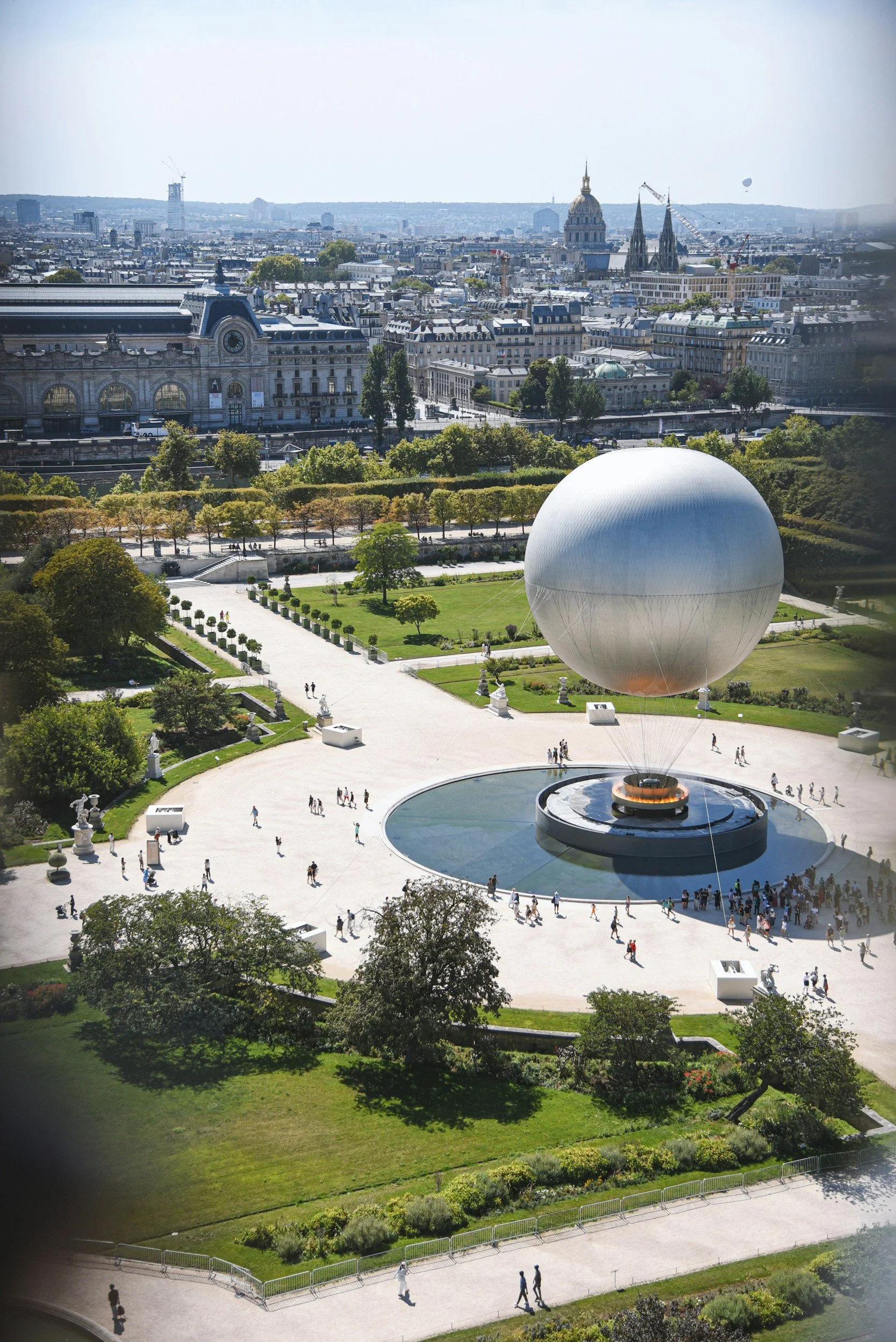 A large white spherical structure suspended above a fountain in a park with green trees and pathways, overlooking a cityscape with historic buildings and spires.
