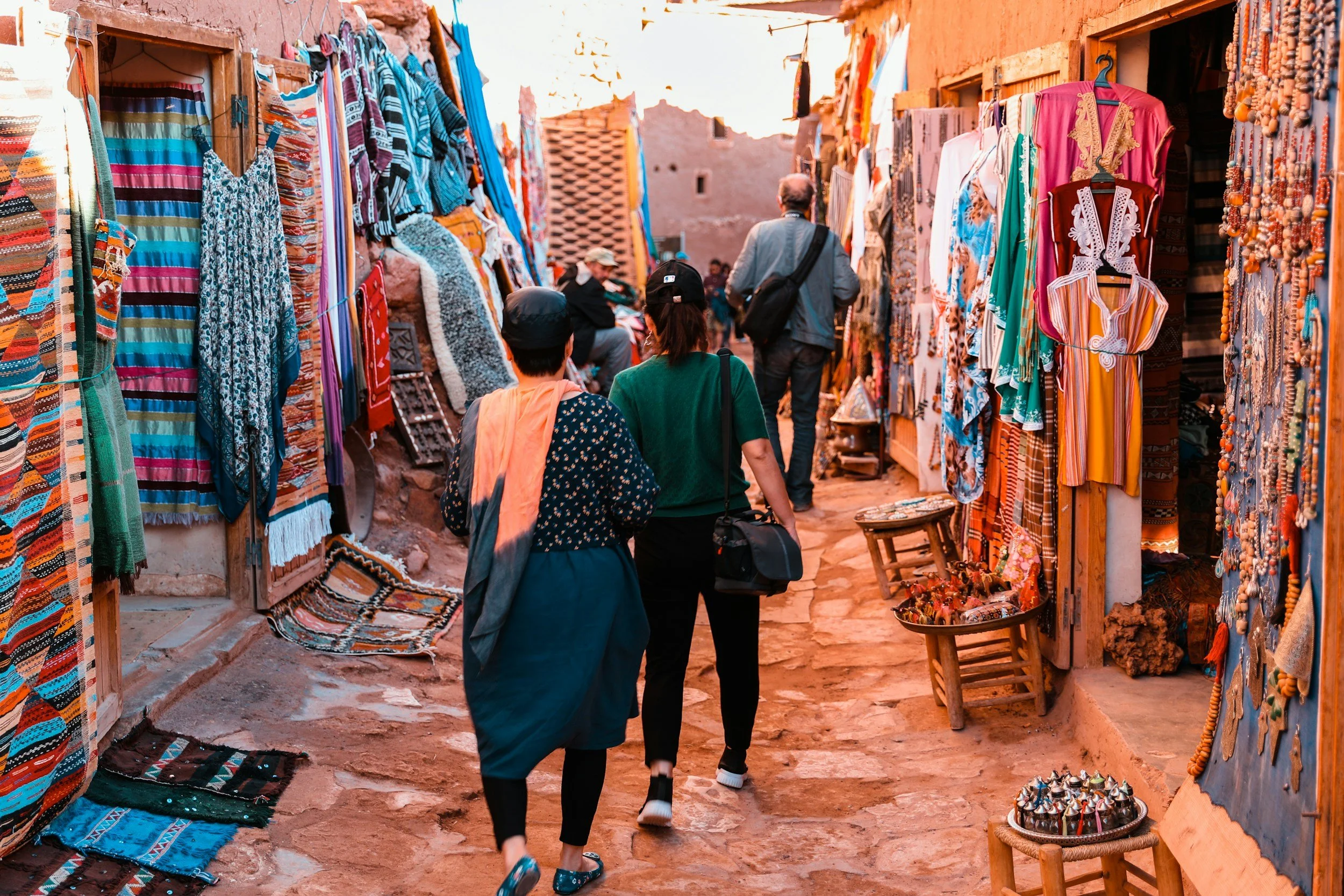 People walking through a colorful marketplace with stalls selling textiles, clothing, and jewelry, with reddish-brown buildings in the background.