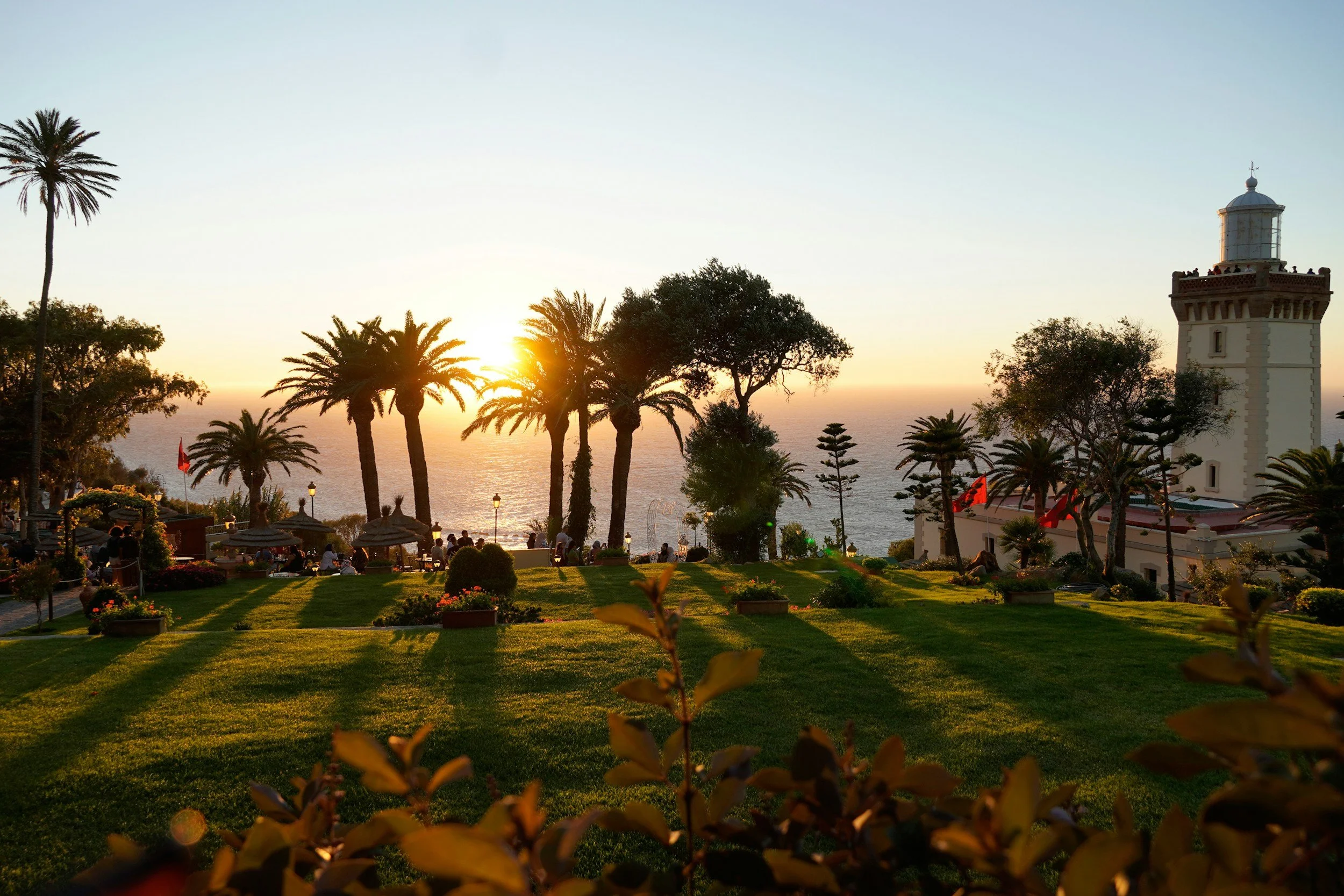 Sunset over a park with palm trees, a lighthouse, and people sitting and walking near the ocean.