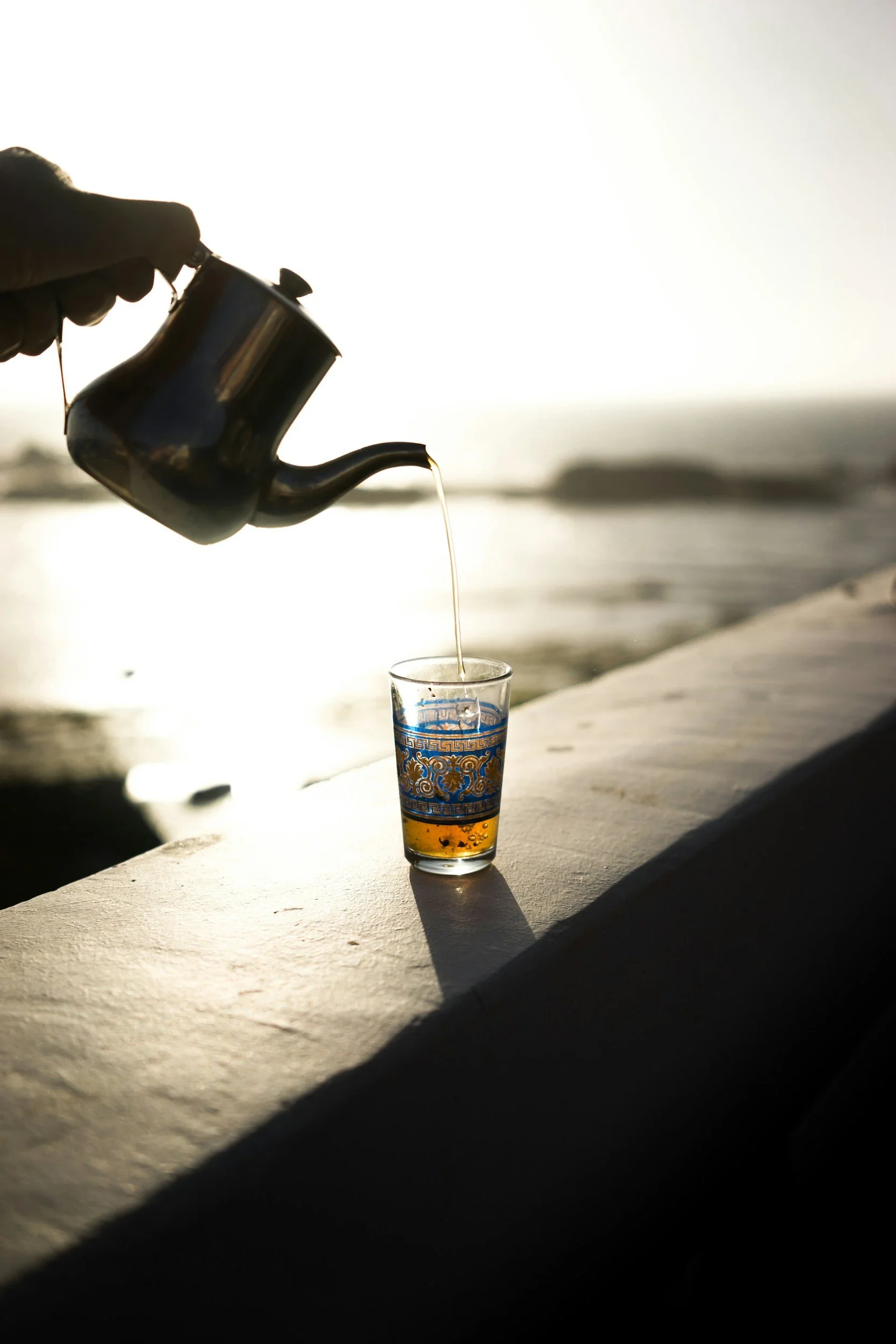 A hand holding a small metallic teapot pouring hot tea into a glass with blue and gold patterns, set on a concrete surface near a body of water during sunset or sunrise.