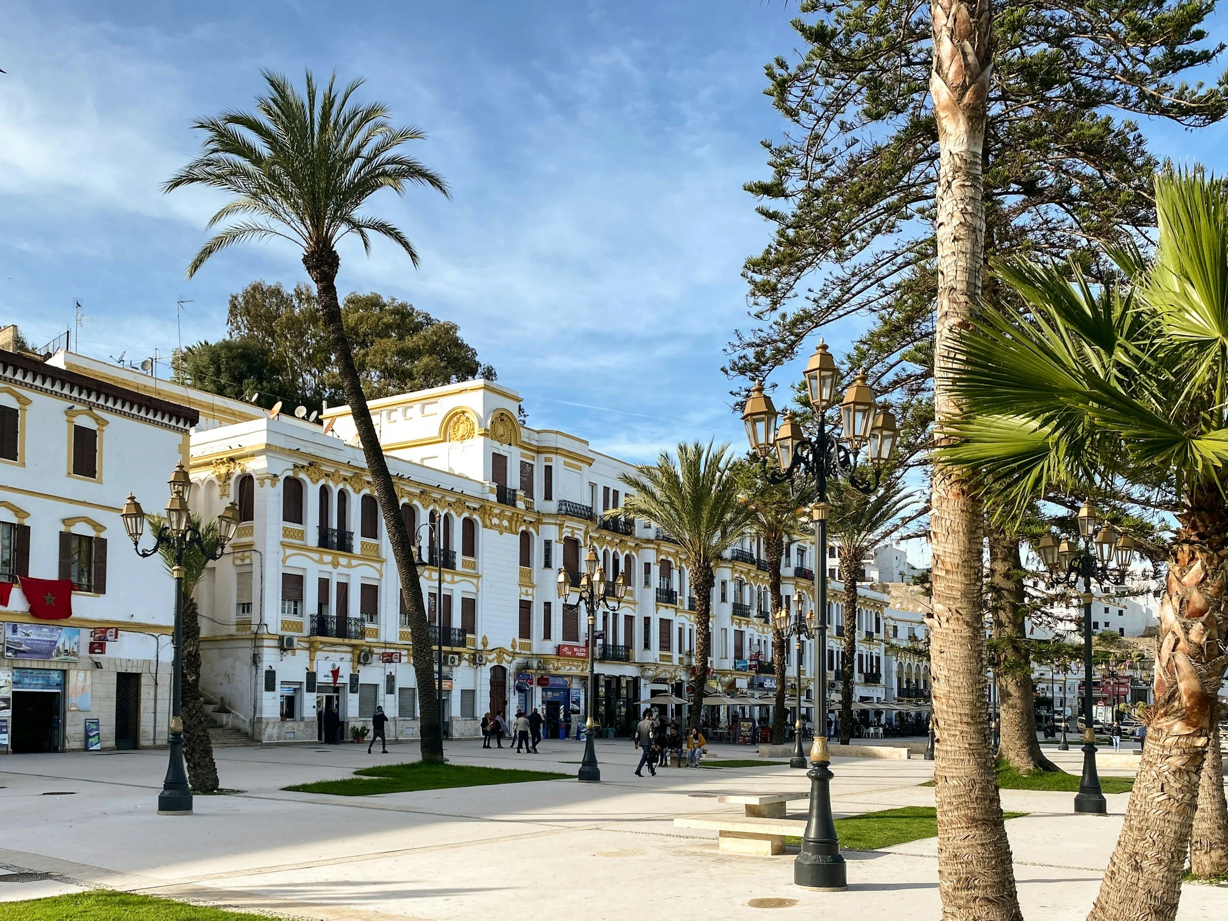 City square with white buildings, palm trees, street lamps, and people walking under a blue sky.