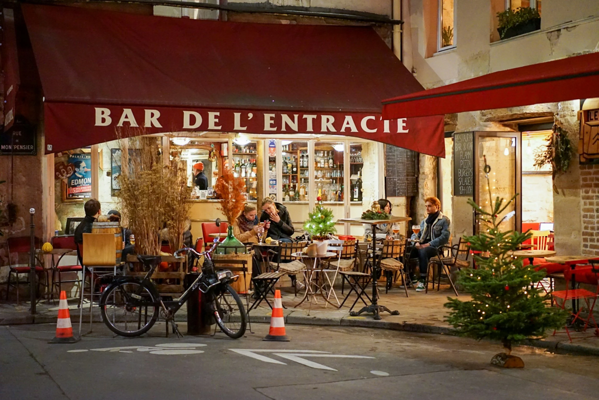 Outdoor seating area of a bar called 'Bar de l'Entracite' at night with a Christmas tree, bicycles, and a few people sitting and chatting outside.