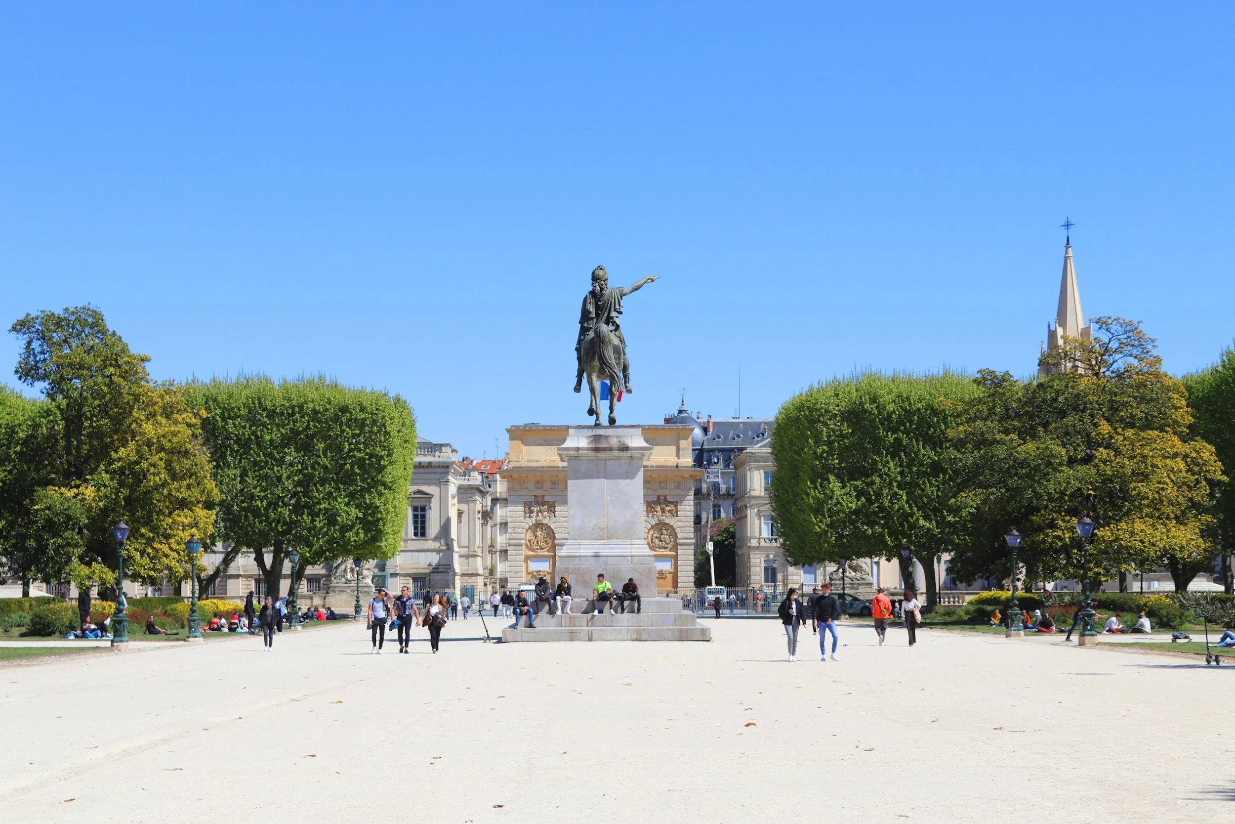 A park with a statue of a man on a horse at the center, surrounded by green trees and people walking or sitting on benches, under a clear blue sky.