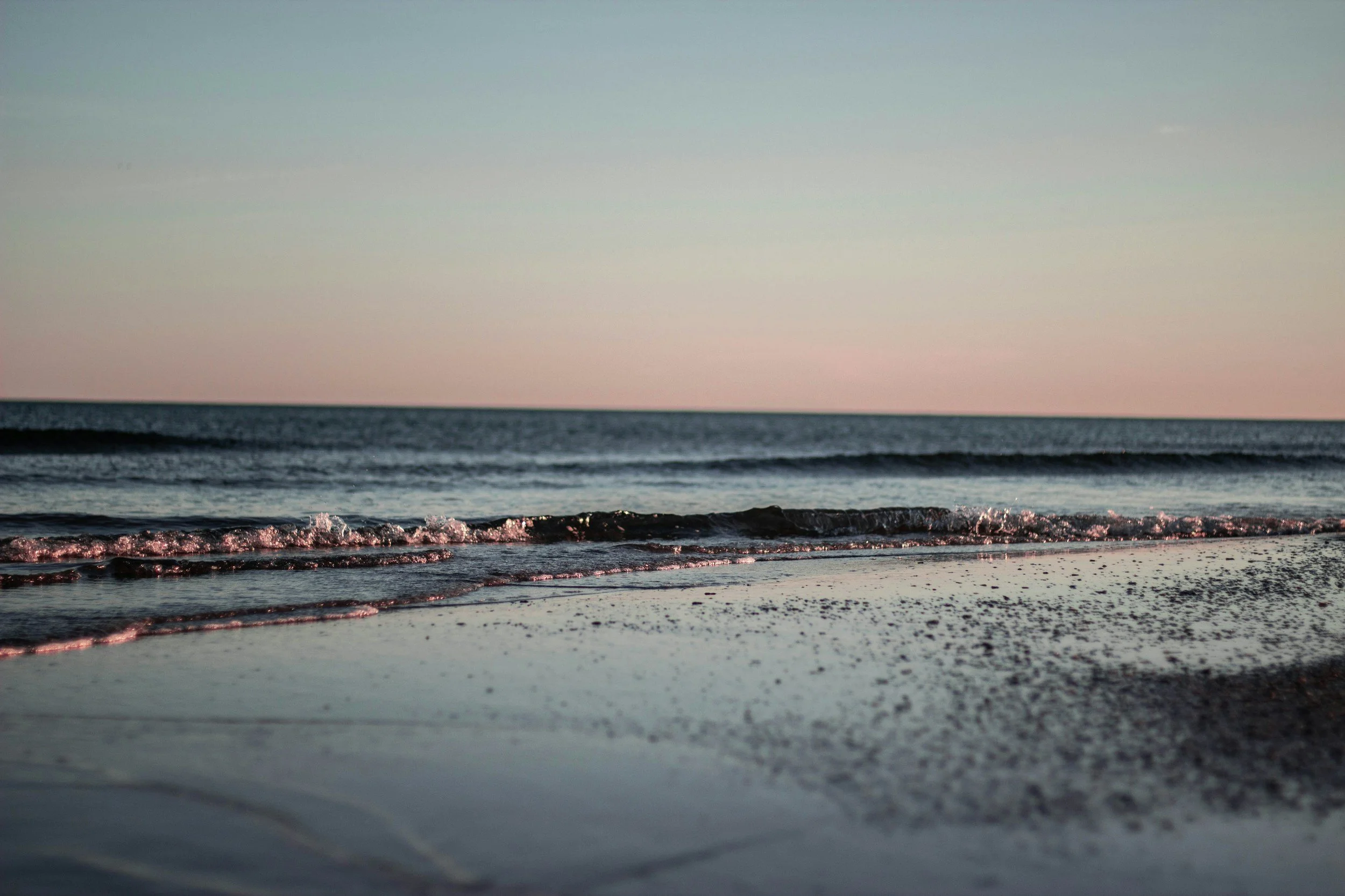 A serene beach scene at sunset or sunrise with gentle waves lapping at the sandy shore, under a clear sky.
