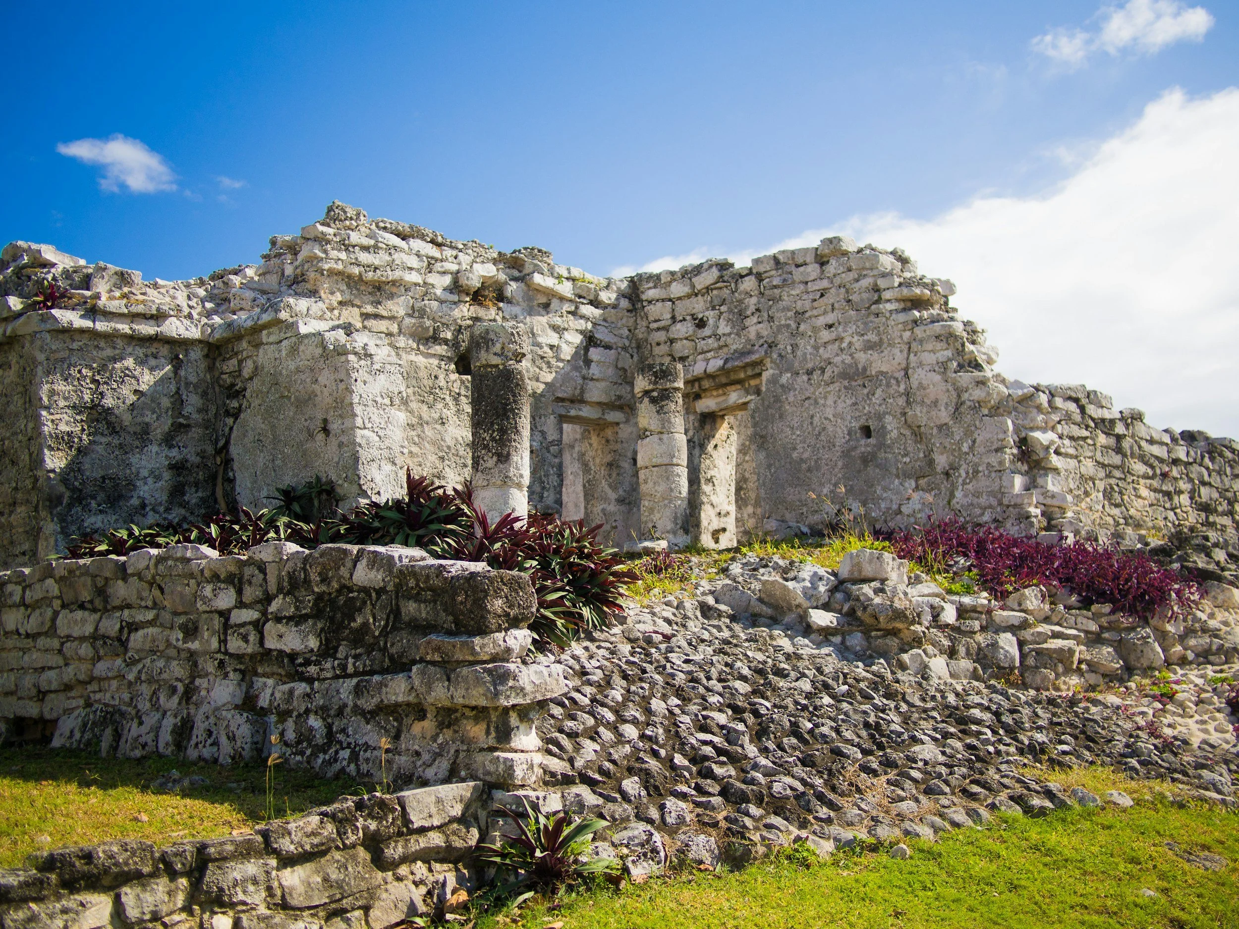 Ruins of an ancient stone structure with small columns, surrounded by greenery and purple plants under a blue sky with clouds.