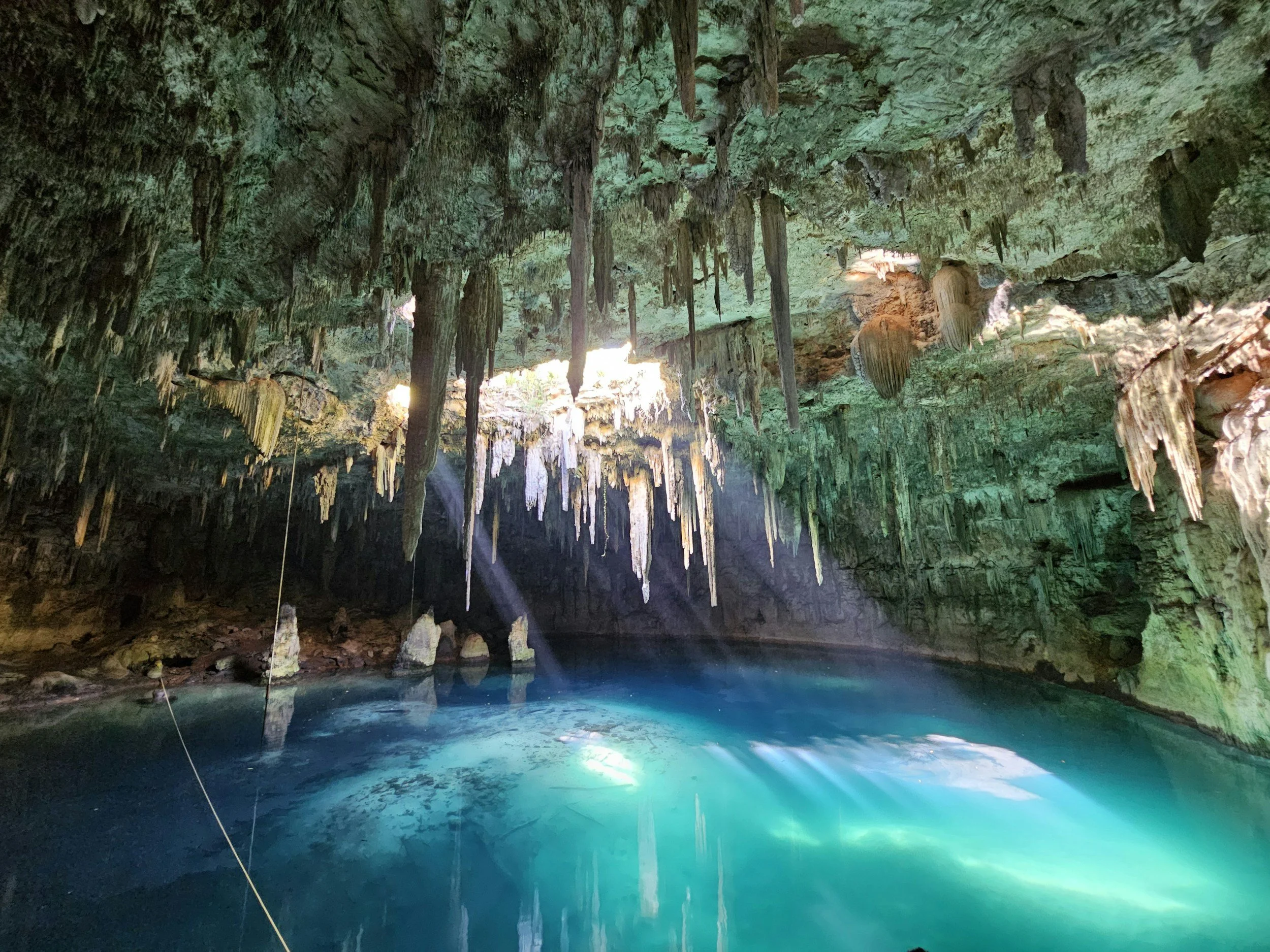 Inside a cave with hanging stalactites and illuminated turquoise water reflecting light.