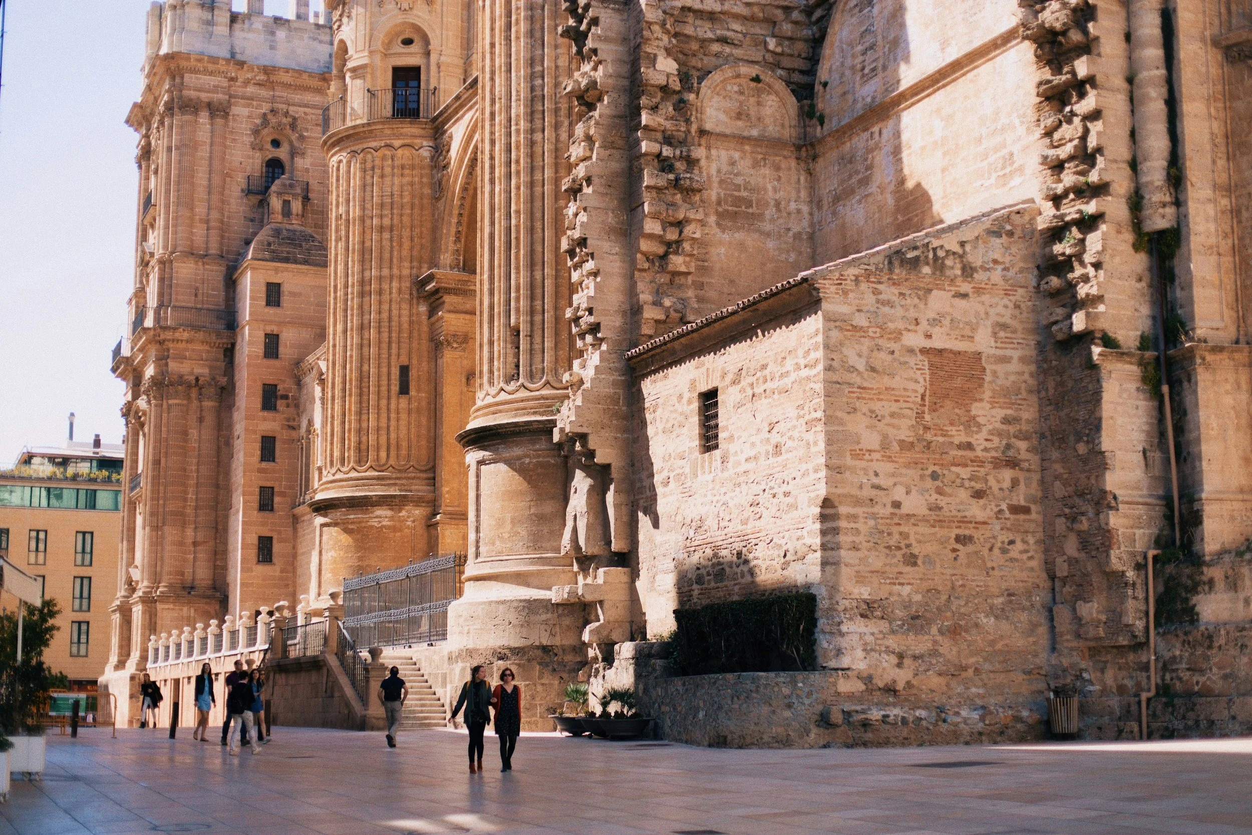 A historic stone building with elaborate architectural details, including columns and carvings, alongside tourists walking on a paved plaza.