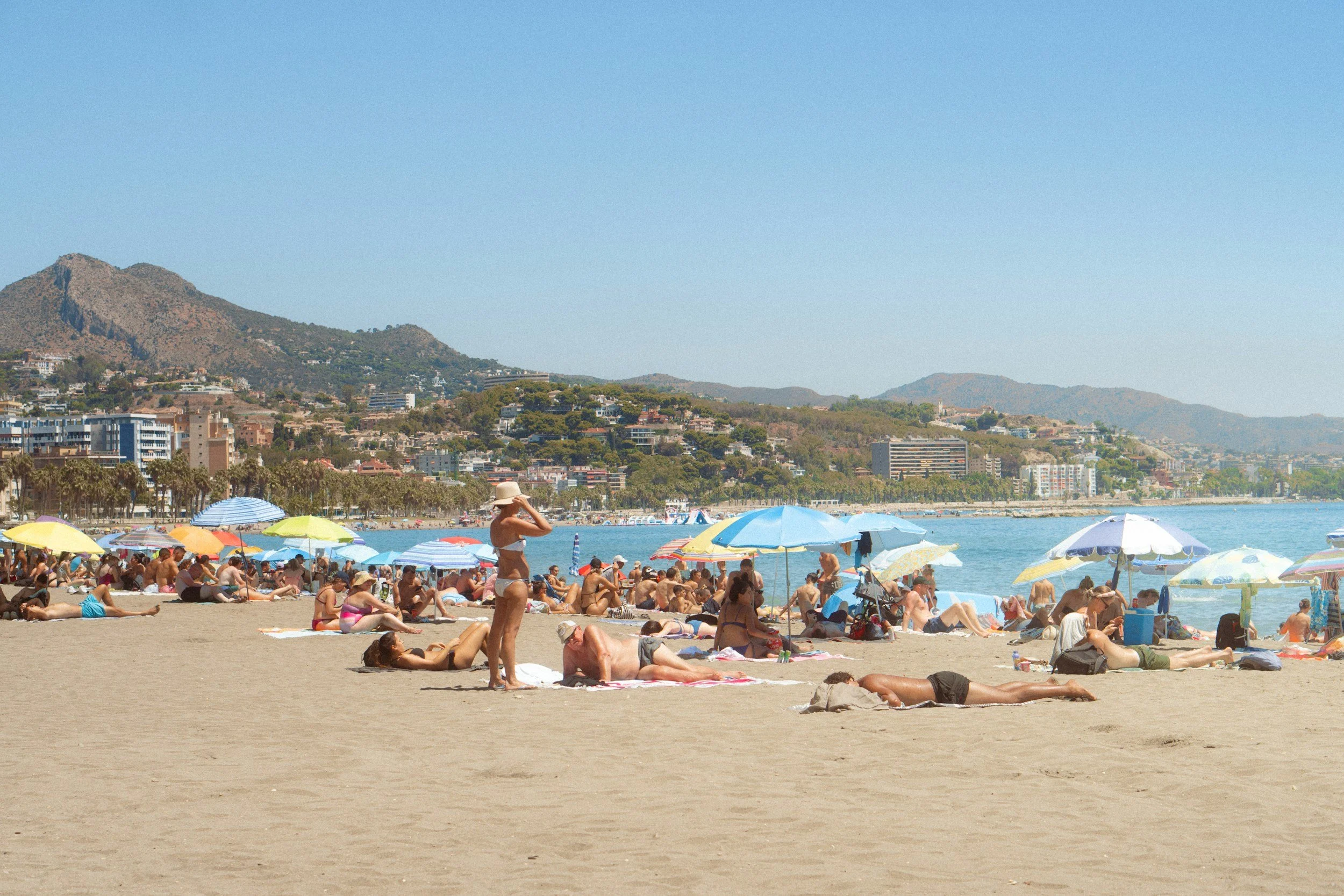 Beach scene with numerous people sunbathing and under umbrellas near the water, with hills and buildings in the background on a clear, sunny day.