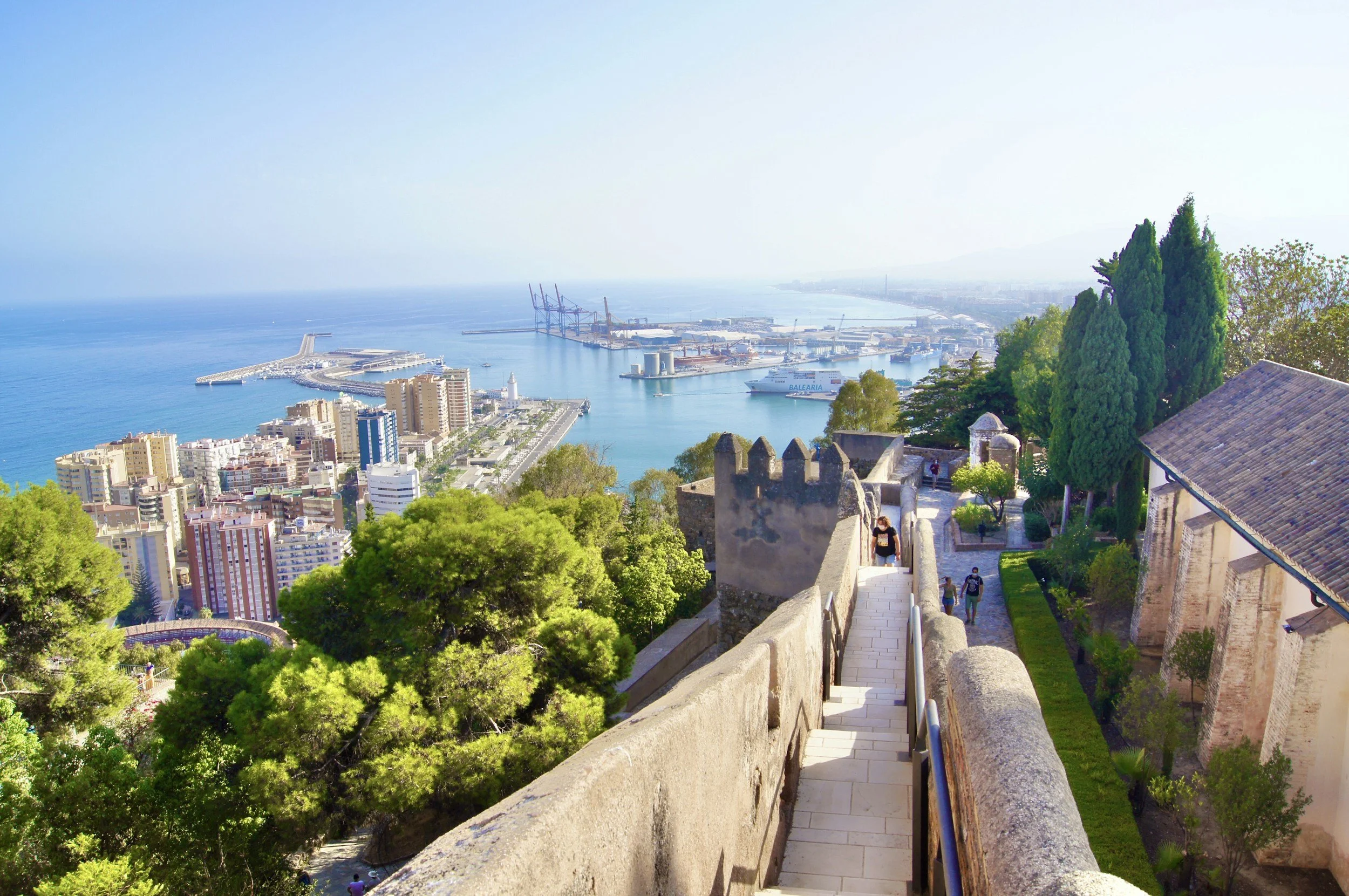 A scenic view of a coastal city with a harbor, modern buildings, green trees, and a historic stone fortress wall with a staircase in the foreground.