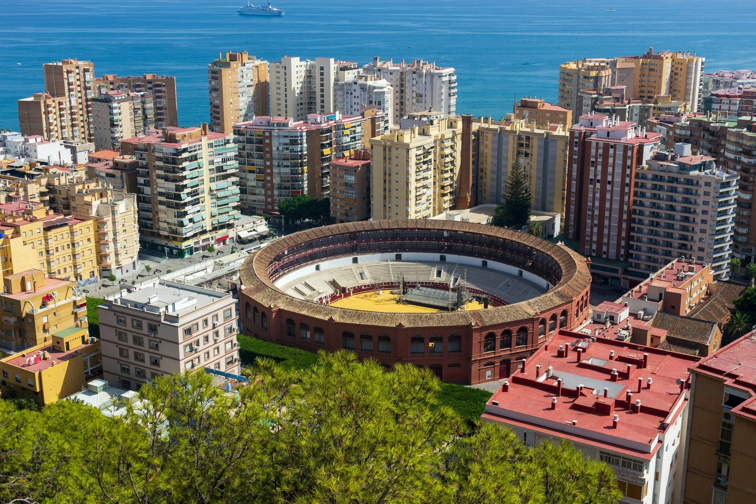 Cityscape with high-rise buildings and a bullring arena in the foreground, overlooking the ocean with a ship in the distance.