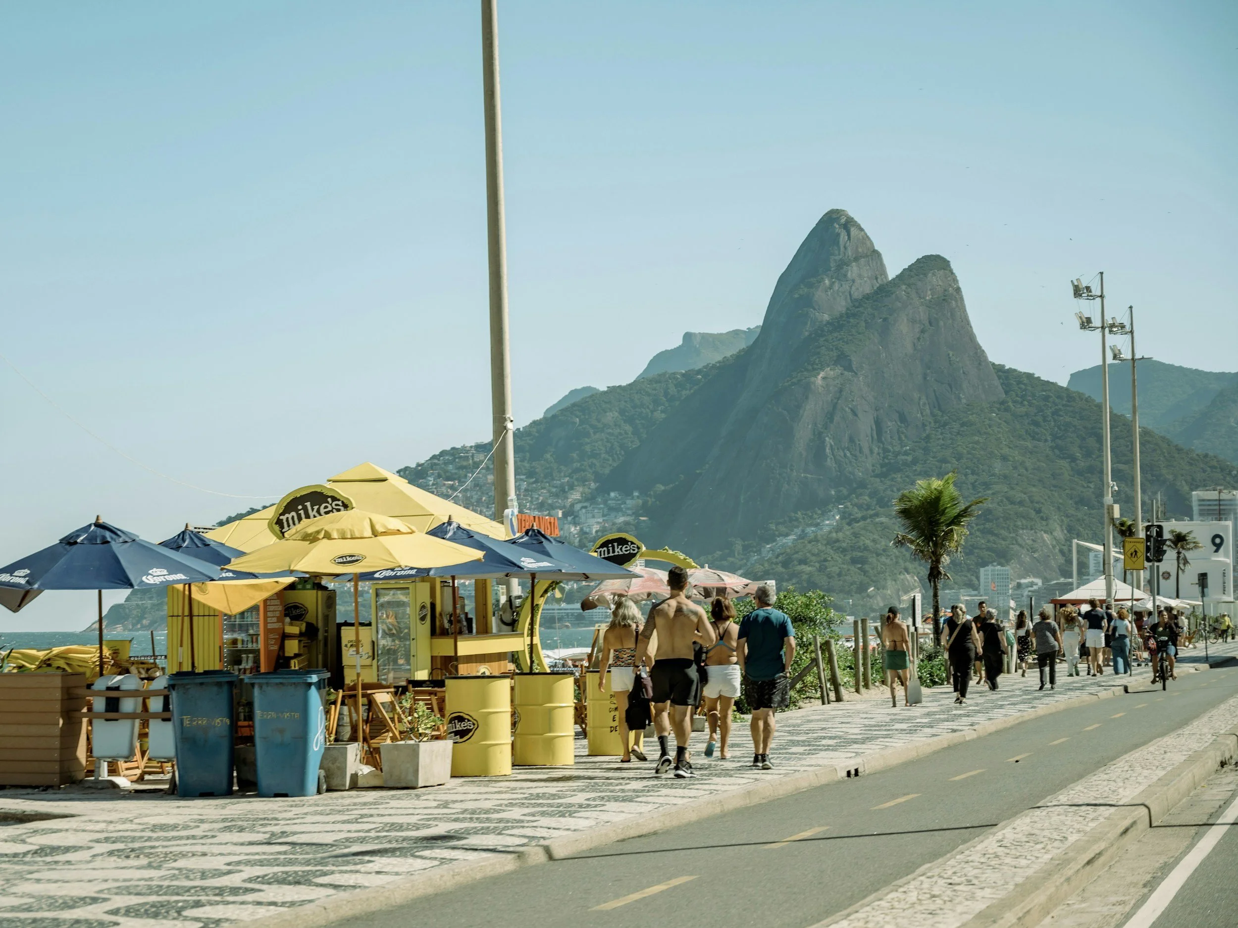 Beachside walkway with vendors and people, Sugarloaf Mountain in the background, clear sky, palm trees and umbrellas.