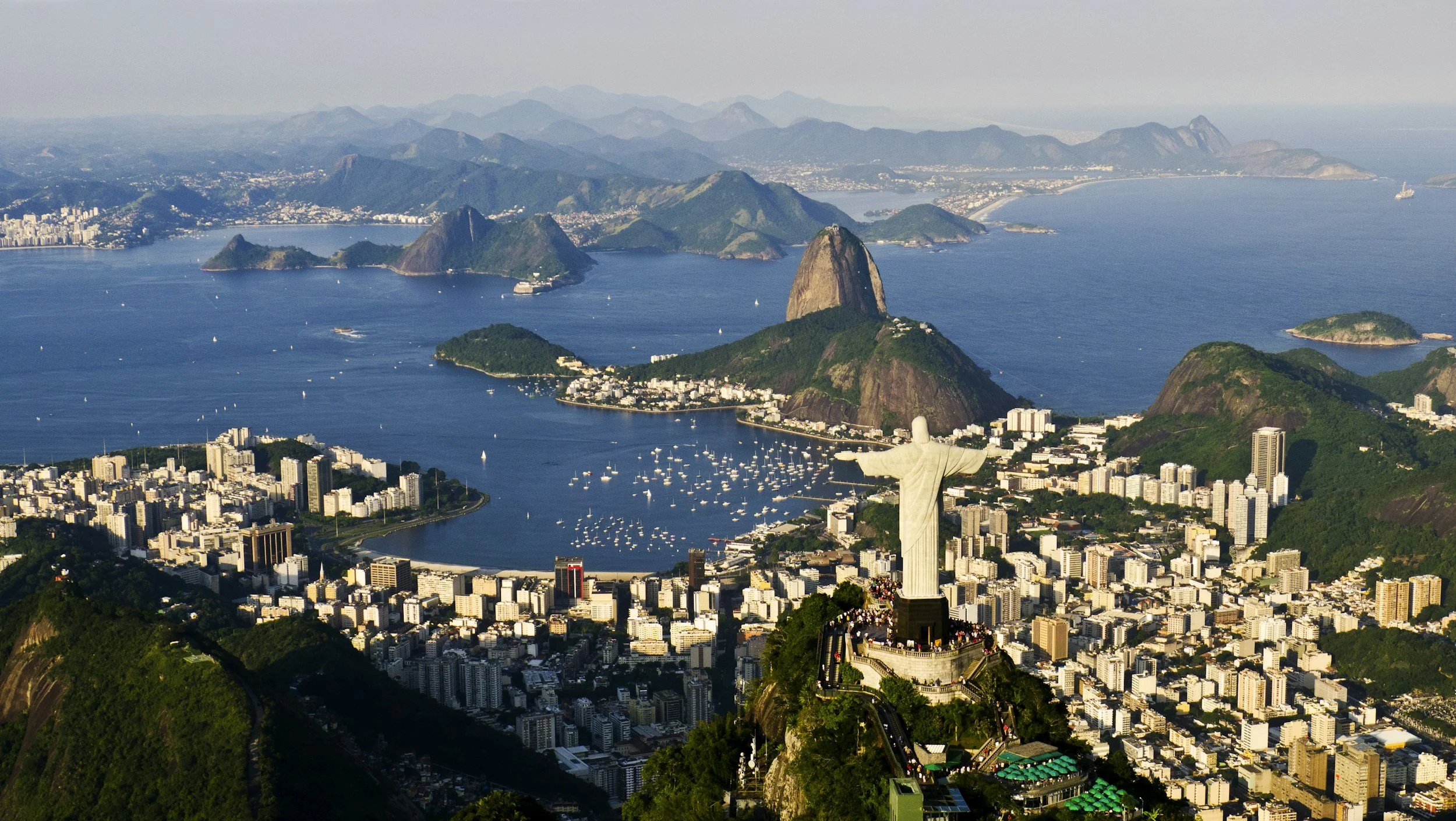Aerial view of Rio de Janeiro featuring Christ the Redeemer statue on Corcovado Mountain overlooking the city, with Sugarloaf Mountain, bays, boats, and surrounding hills.