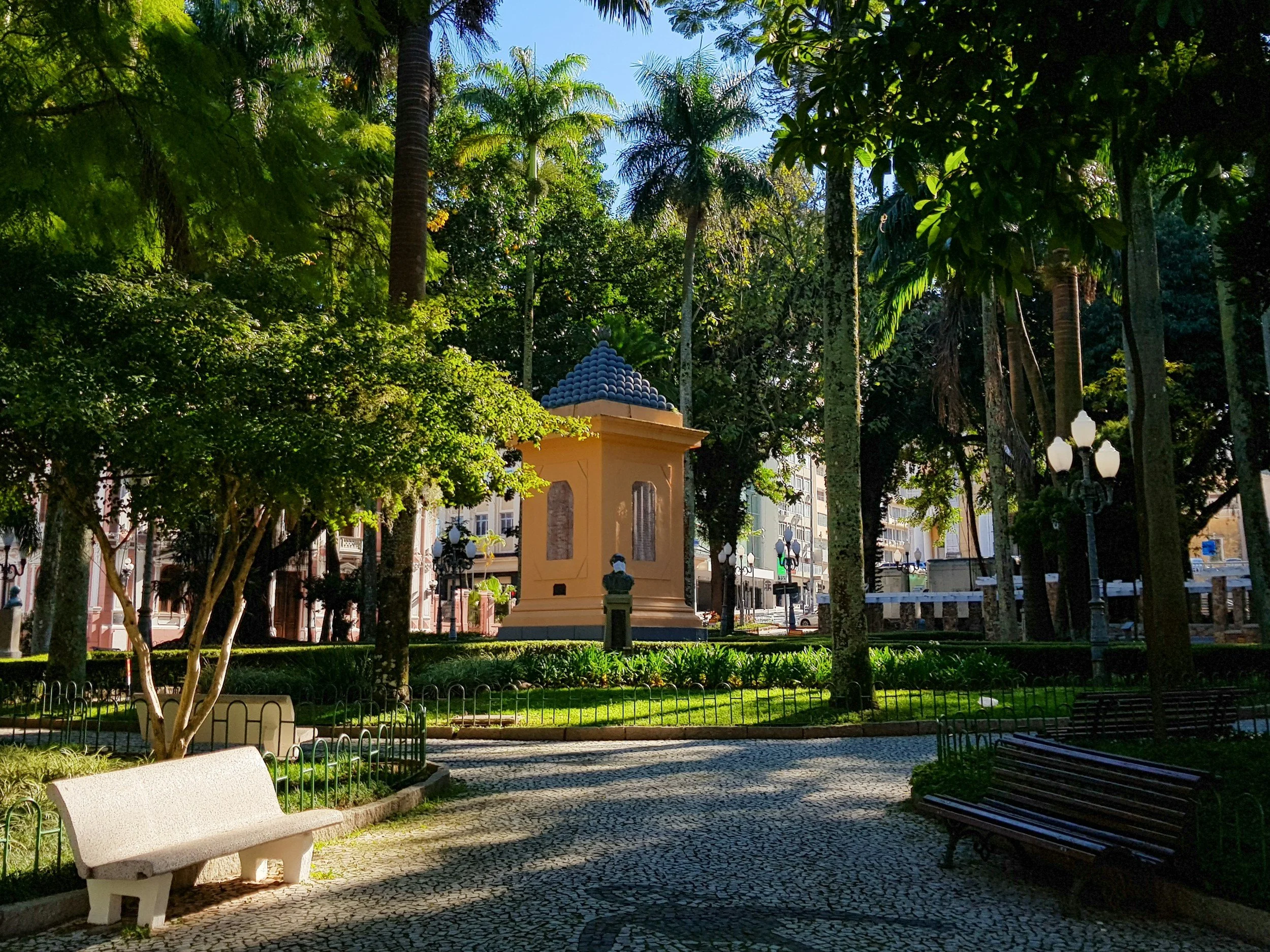 A park with benches, trees, and a small structure with a blue-tiled roof, surrounded by lamp posts and buildings in the background.