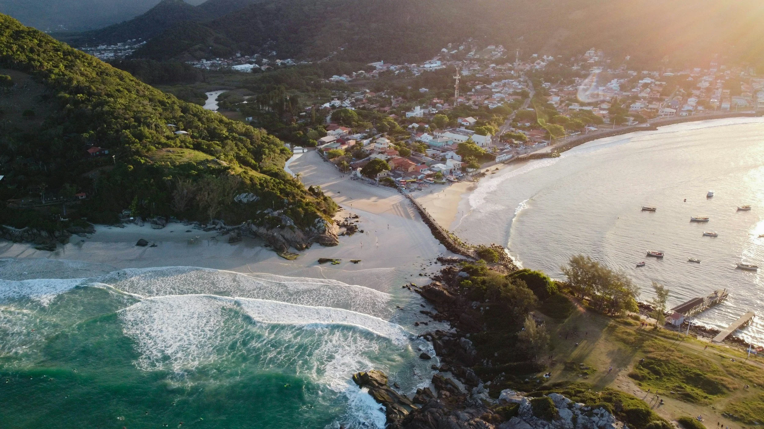 Aerial view of a coastal town with a sandy beach, turquoise ocean with waves, green hills, and boats in the water during sunset.