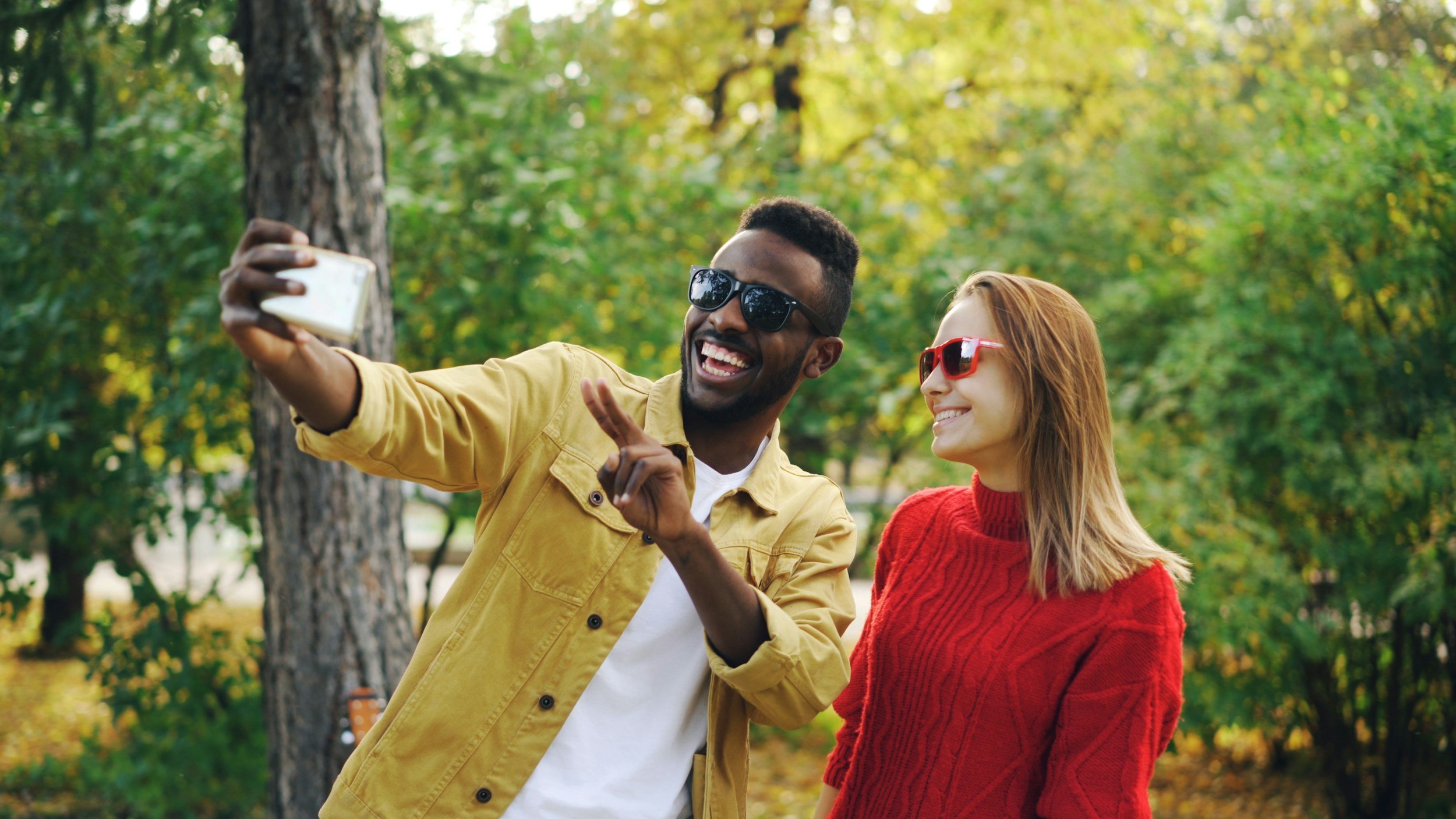 A man and woman standing outdoors taking a selfie with a smartphone, both smiling and wearing sunglasses, surrounded by green foliage and trees.