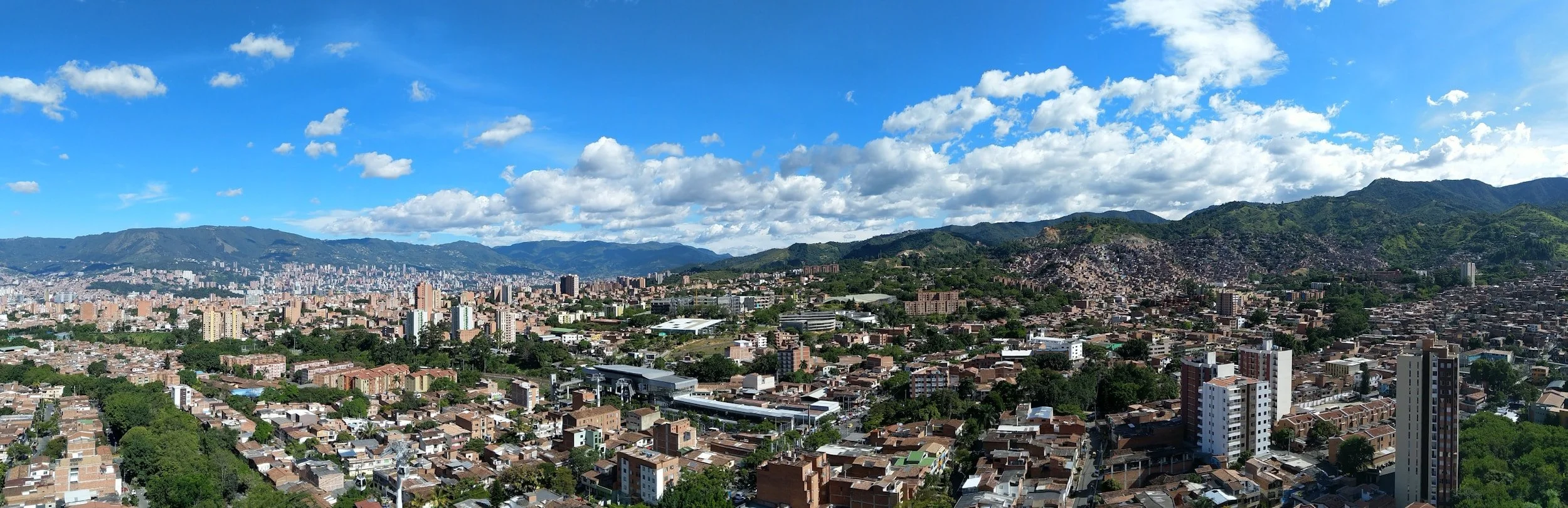 Panoramic view of a city with numerous buildings, surrounded by green hills and mountains under a partly cloudy blue sky.