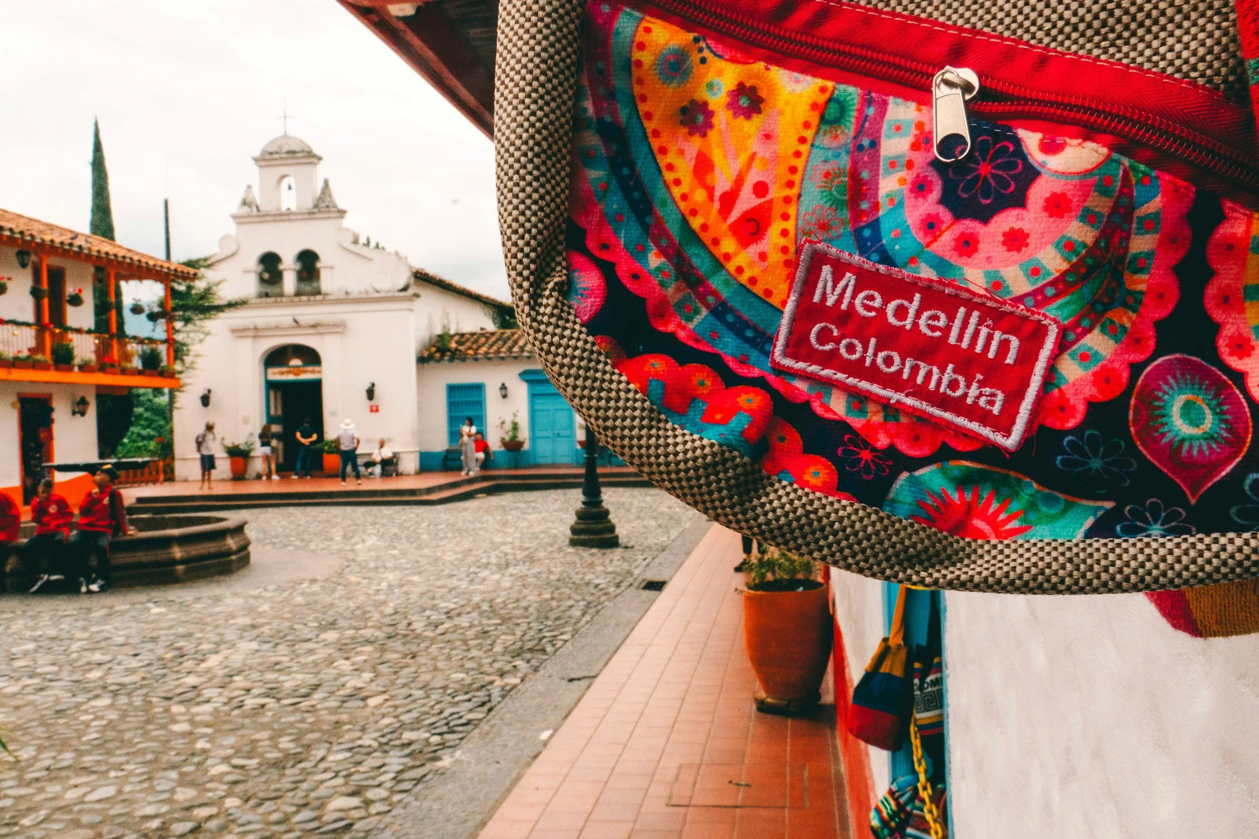 Colorful embroidered backpack with a Medellín, Colombia patch in the foreground, and a colonial-style town square with a white church and cobblestone street in the background.