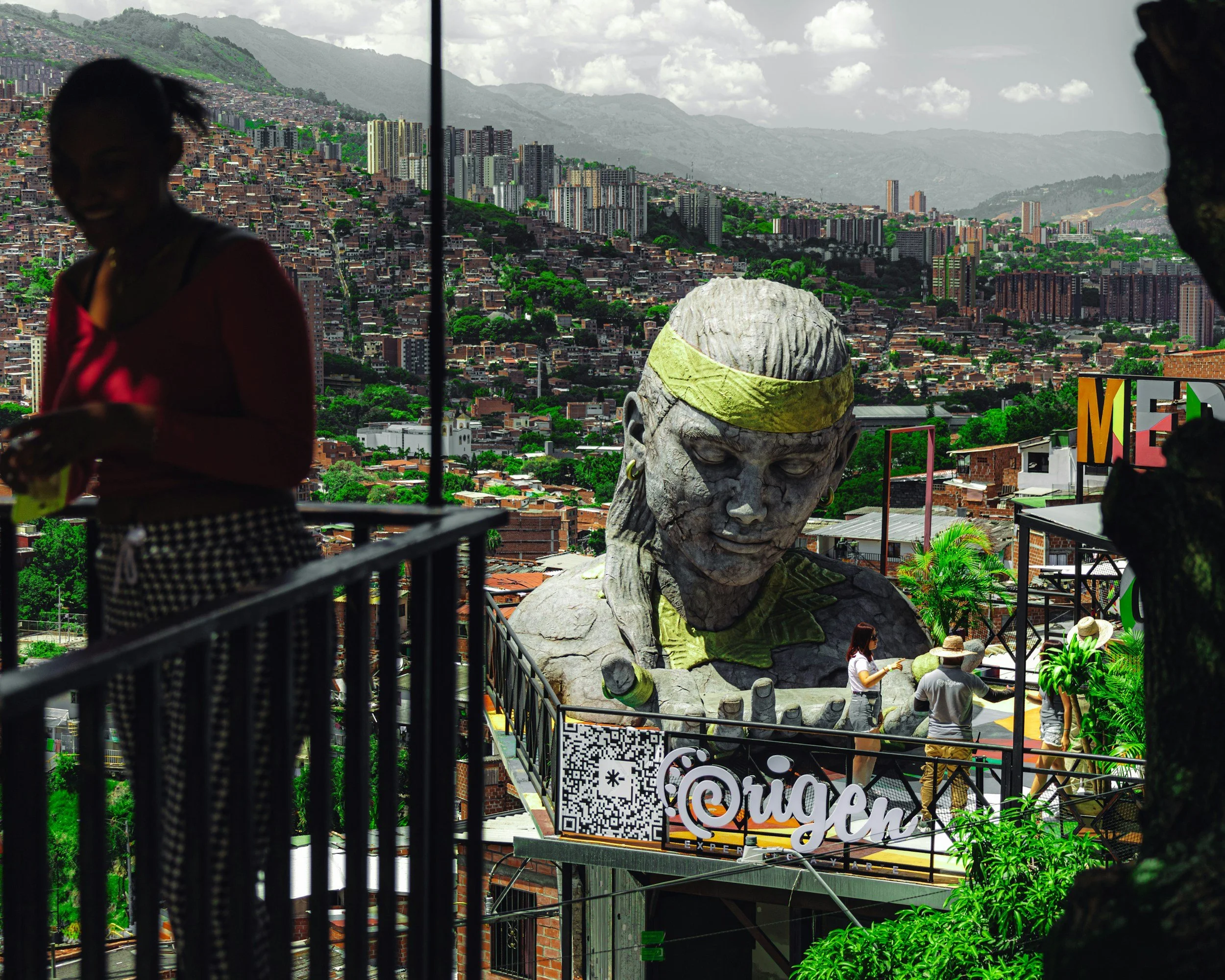 A large outdoor sculpture of a woman's head and shoulders with a yellow headband, surrounded by people, on a balcony overlooking a densely populated hillside cityscape with numerous high-rise buildings and green hills in the background.