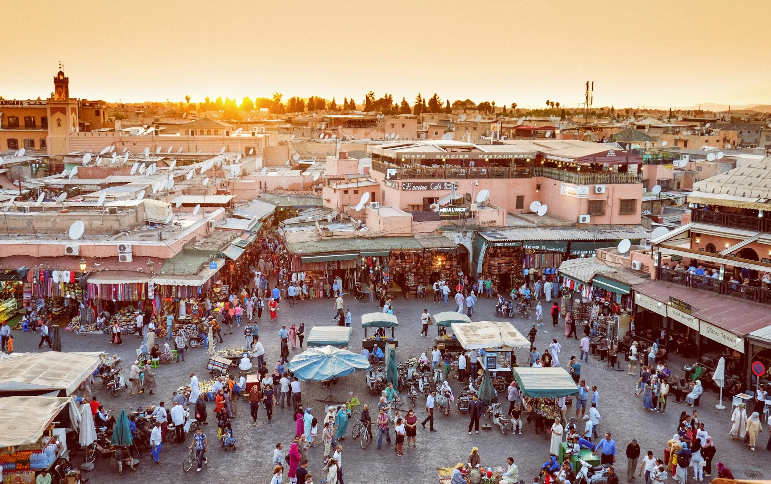 A busy marketplace square in Morocco during sunset, filled with people, market stalls, and vendors, with buildings and satellite dishes in the background.