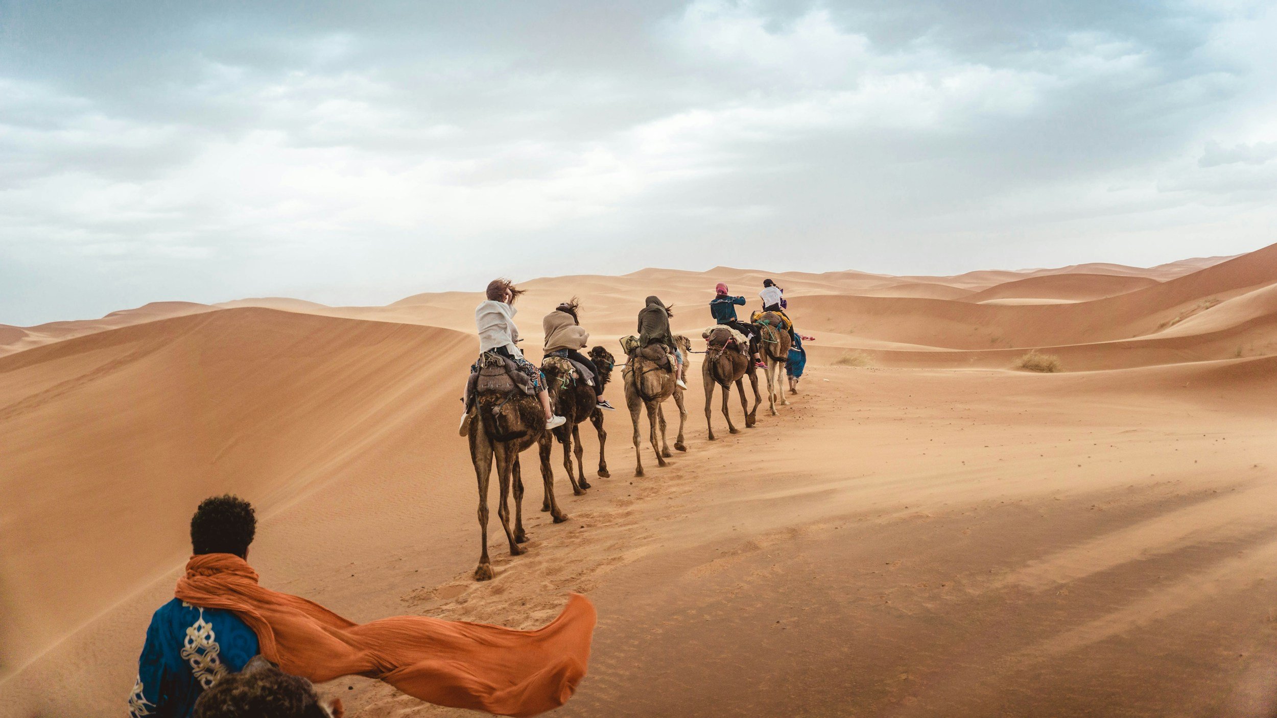 A group of people riding camels through a desert landscape with sandy dunes and cloudy sky.