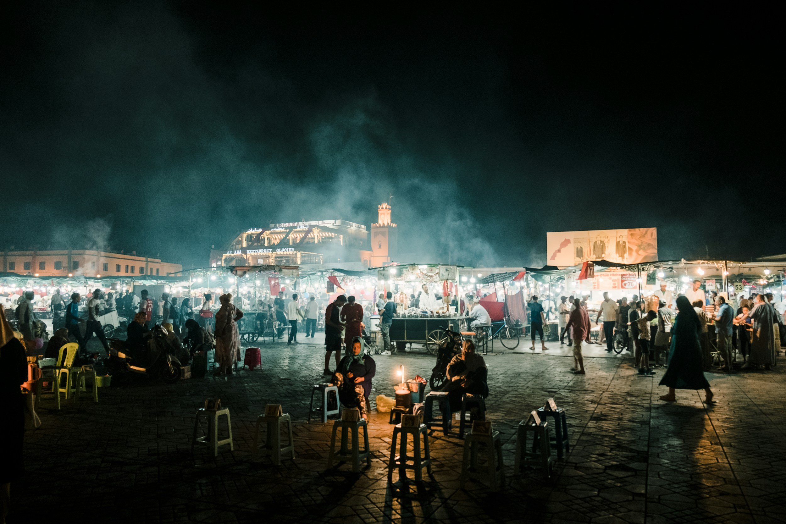 Night market scene with numerous stalls, people, and two women sitting around a small lit candle in the foreground.