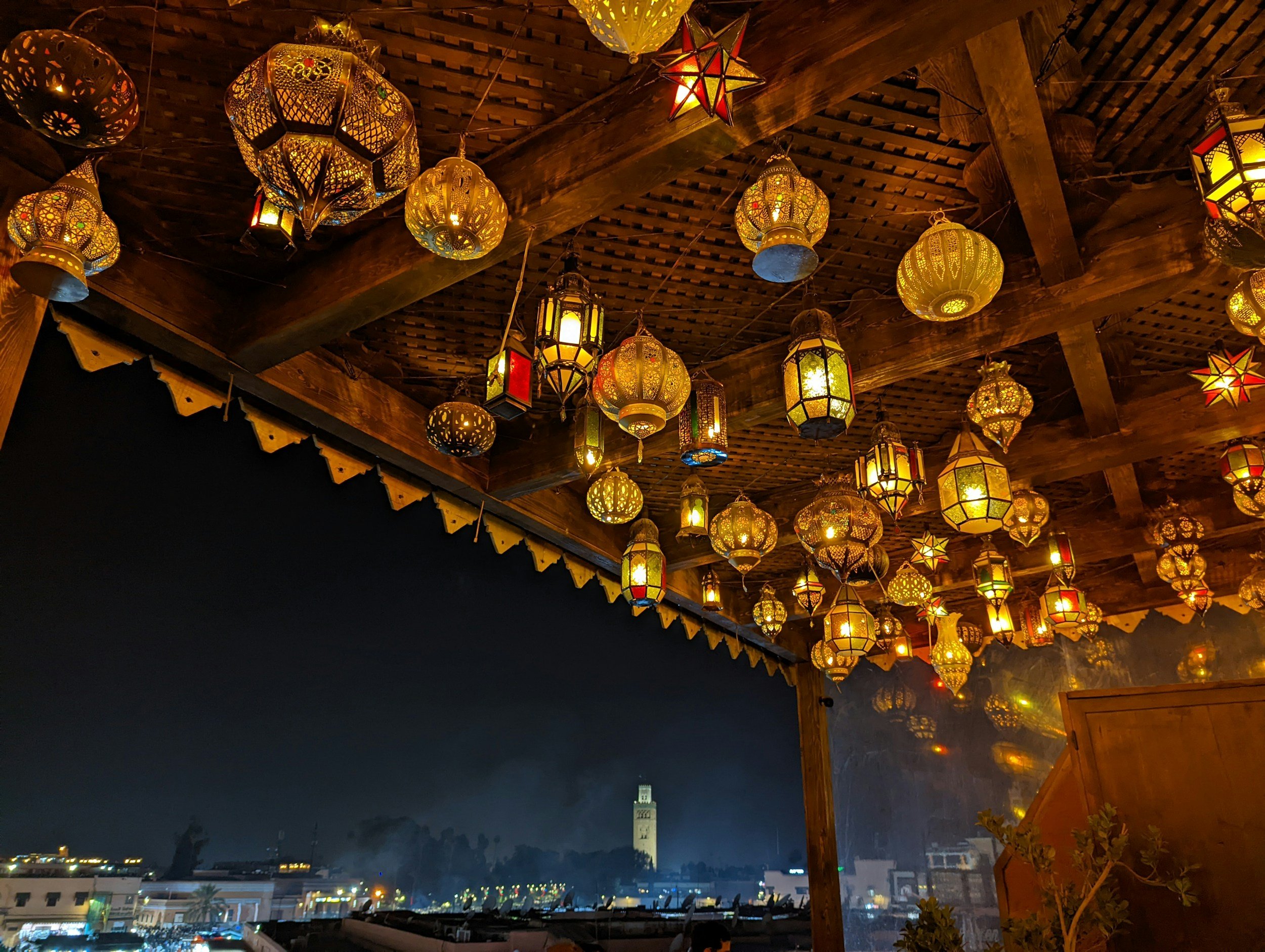Night view of a city skyline with a tall clock tower in the distance, seen from a terrace decorated with numerous colorful Moroccan-style hanging lanterns under a wooden roof.