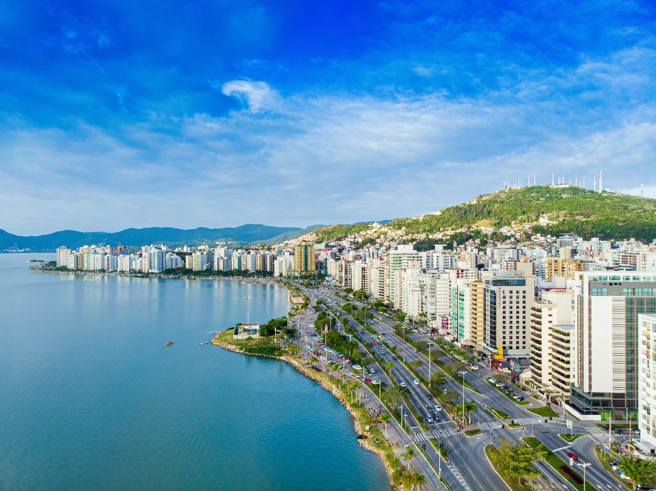 A cityscape along a large bay with numerous high-rise buildings, a road with cars, and a hillside with communication towers, under a partly cloudy sky.
