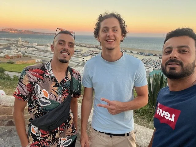 Three men taking a selfie outdoors with a coastal cityscape in the background at sunset.