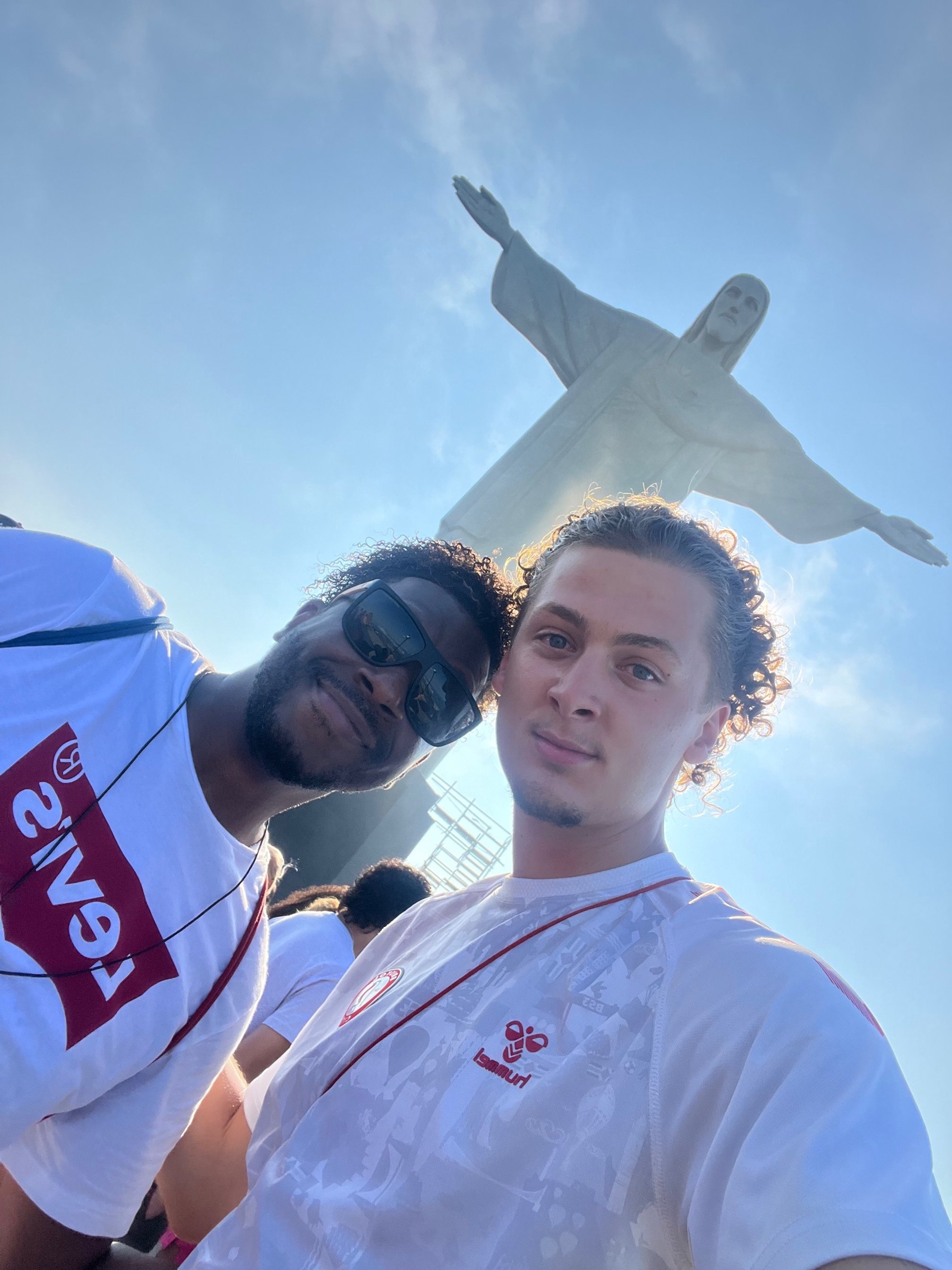Two young men taking a selfie in front of the Christ the Redeemer statue in Rio de Janeiro, Brazil, with clear blue sky background.