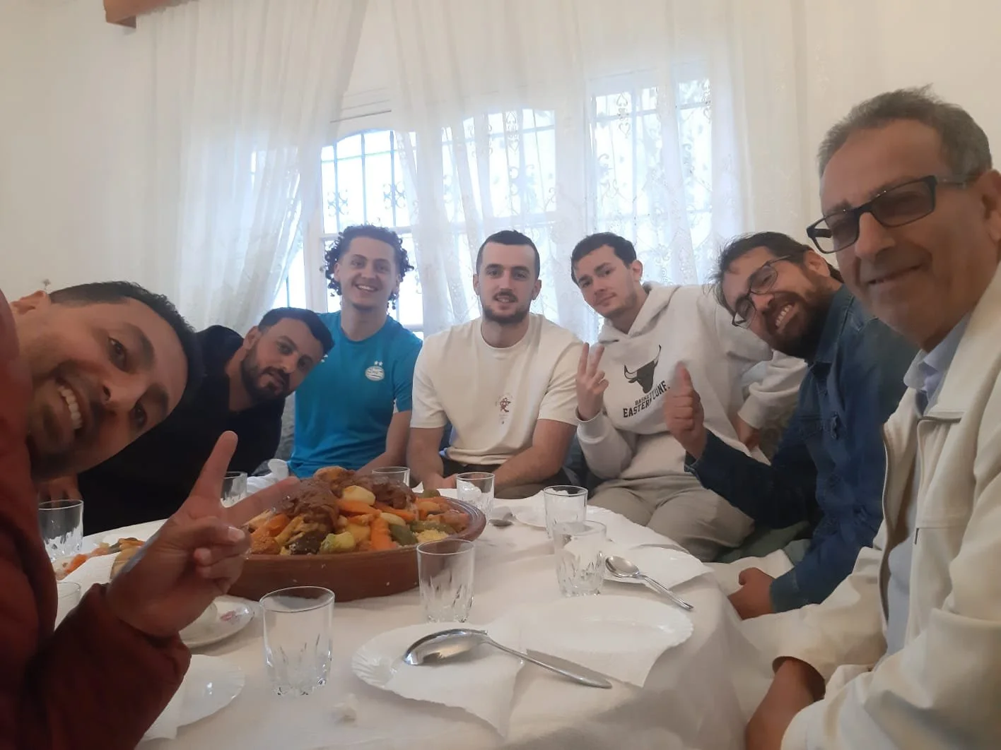 A group of seven men sitting around a table with a large dish of food, smiling and making gestures in a well-lit room with curtains and windows in the background.
