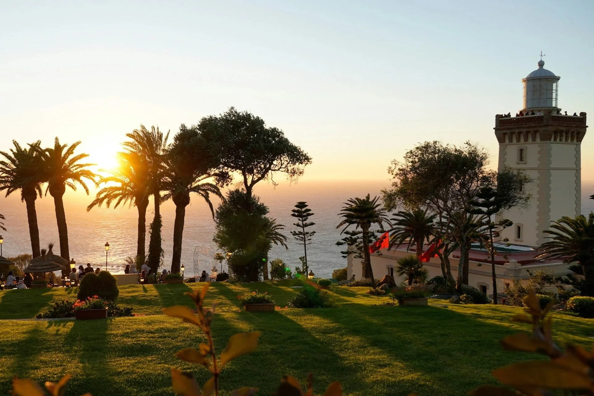 Sunset over a coastal park with palm trees, a lighthouse, and people walking or sitting on benches.