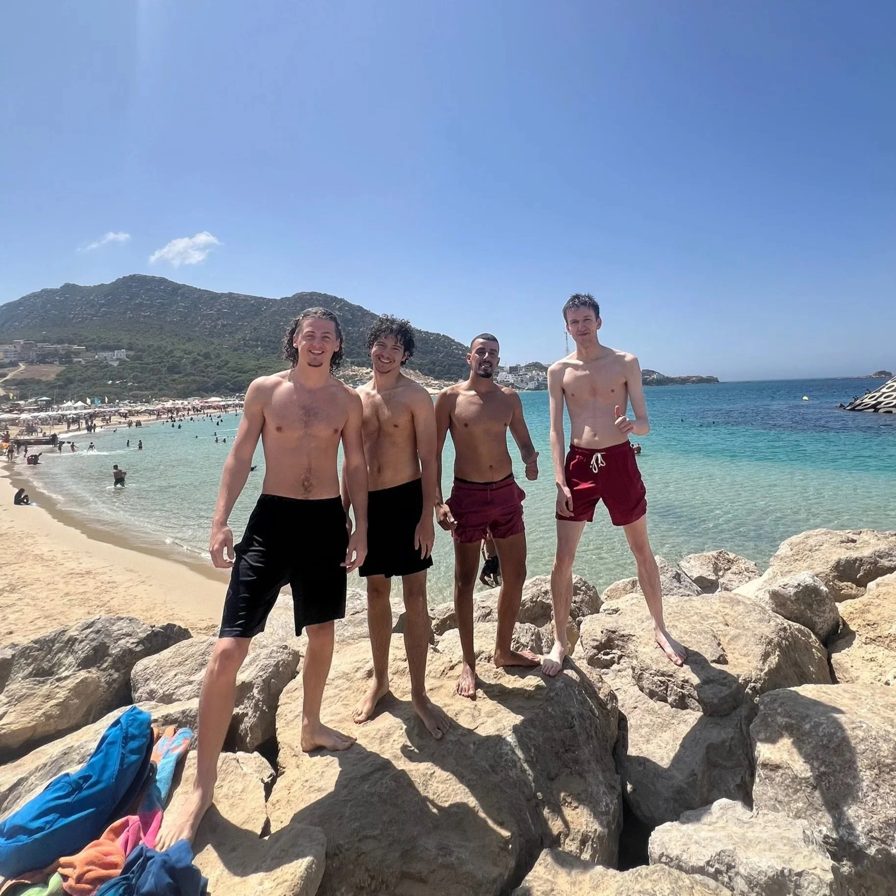 Four young men standing on rocks at the beach, wearing swim trunks, smiling at the camera, with a sandy beach, ocean, and hills in the background.