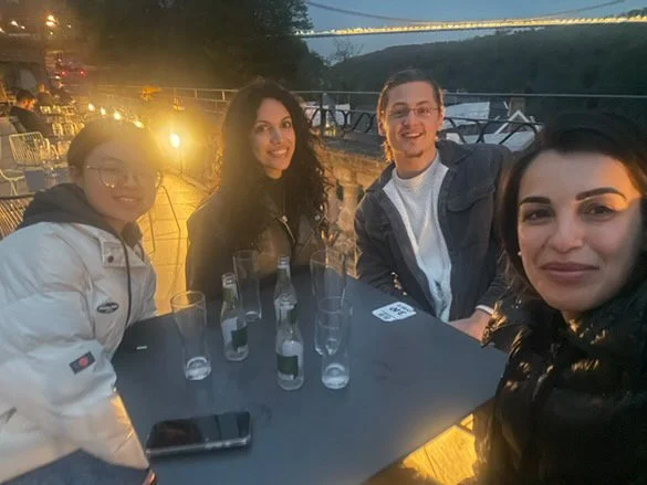 Four people sitting at a table outdoors during the evening, with a scenic view and a bridge in the background. They are smiling, with empty glasses and bottles on the table.