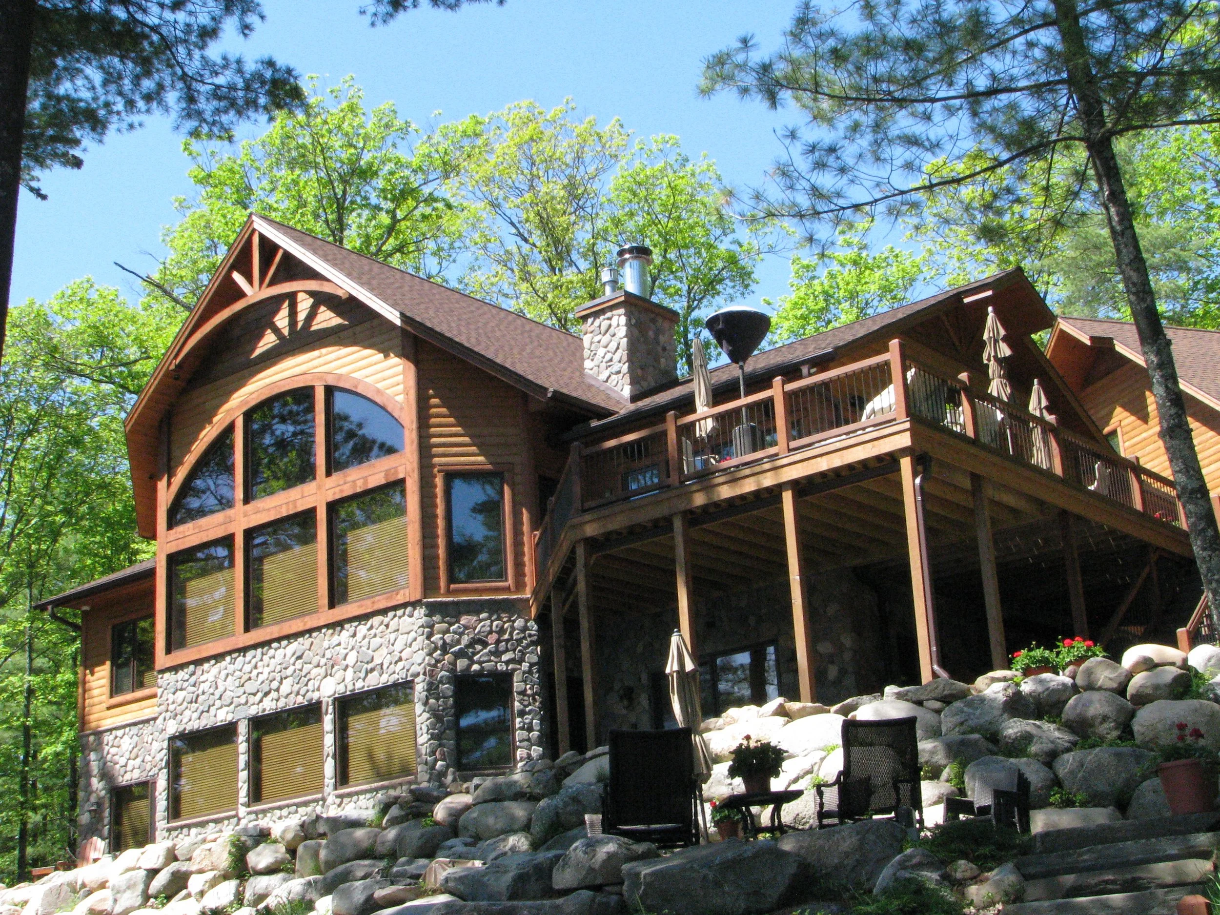 Wooden house on a rocky hill with large trees surrounding it and a clear blue sky above.