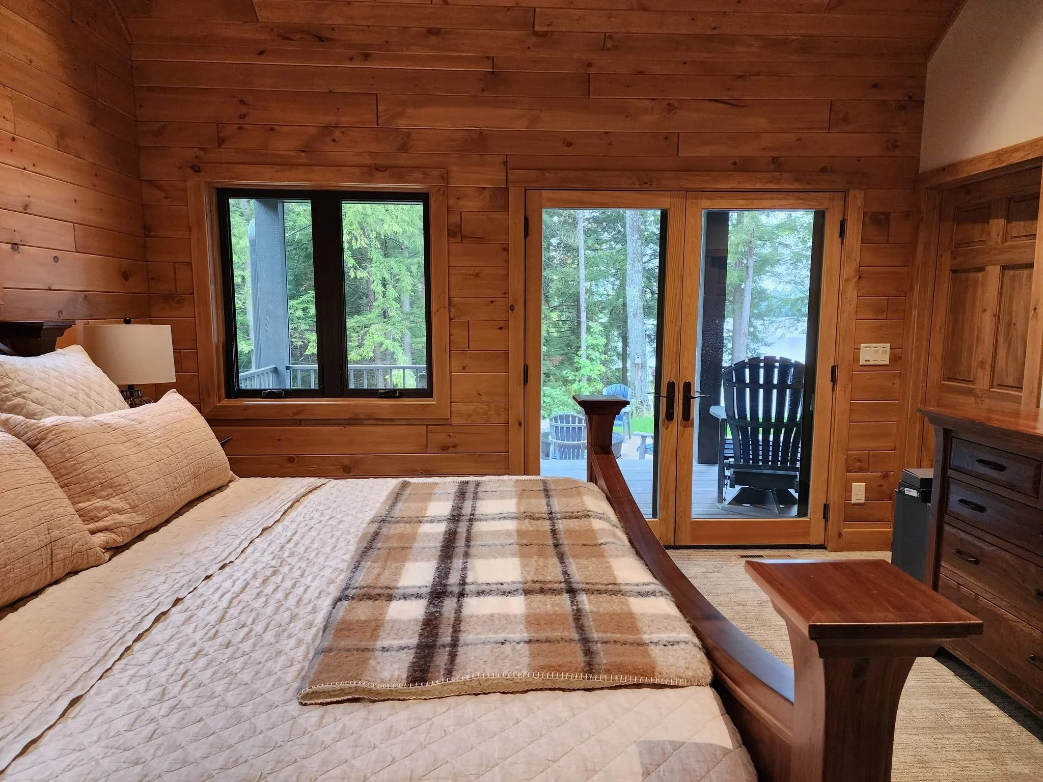 Bedroom with wooden walls, a bed with beige pillows and a plaid blanket, a window and glass sliding door opening to a deck with outdoor chairs, surrounded by trees.