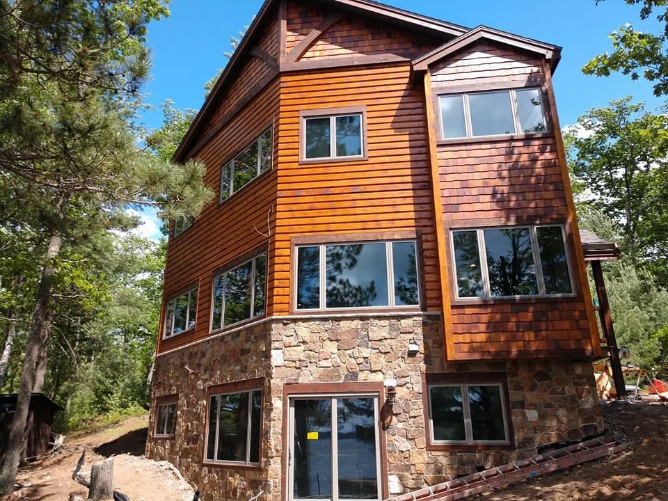 A multi-story house with a stone foundation and wooden upper floors, surrounded by trees under a clear blue sky.