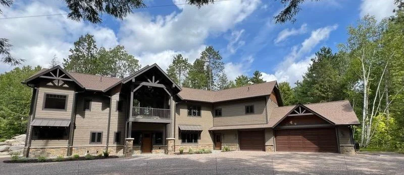 Large wood-style house with multiple gables, surrounded by trees, with a gravel driveway in front.