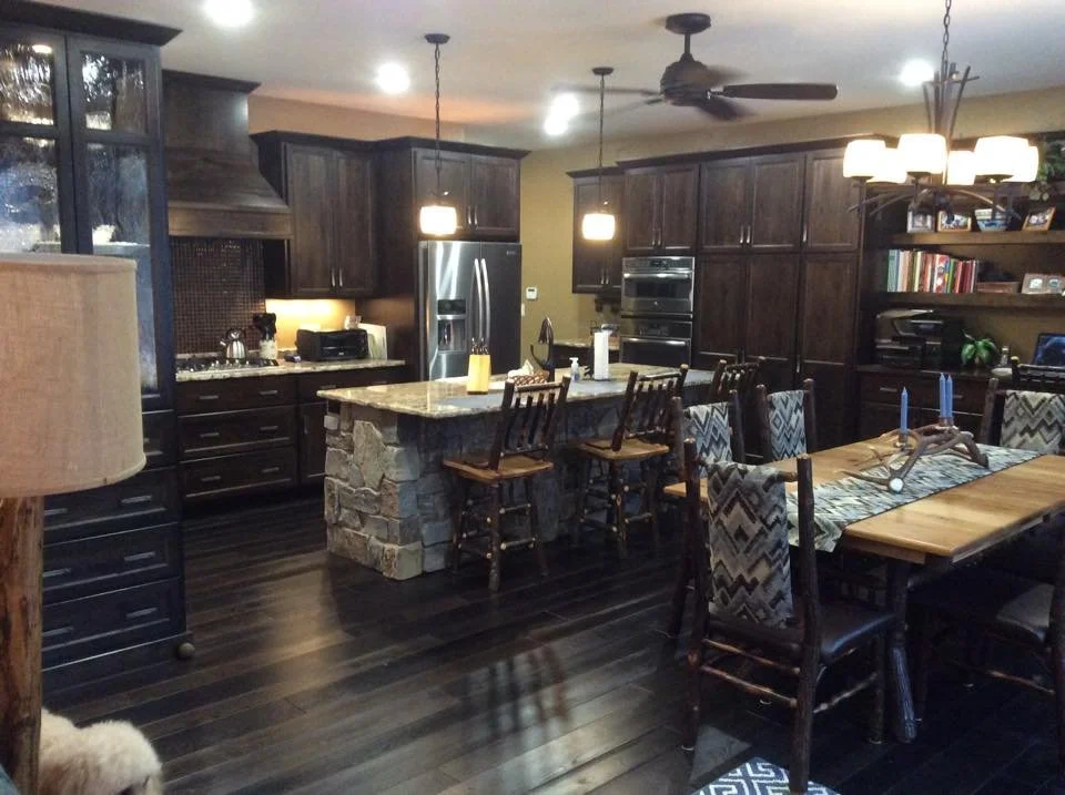Kitchen and dining area with dark wood cabinets, a stone island, stainless steel fridge, wooden dining table with patterned chairs, hanging lights, and a bookshelf.