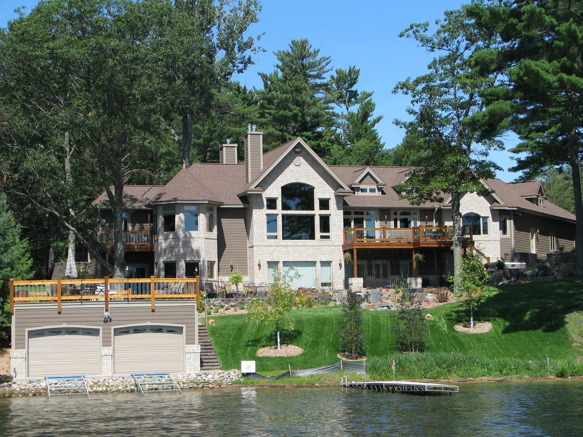 Large house with multiple levels and a mix of brick and brown siding, featuring multiple decks and large windows, situated on a lush green lawn, by a body of water with a dock in the foreground, surrounded by tall trees.