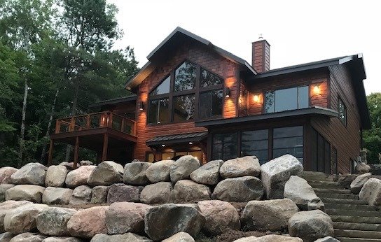 A modern two-story wooden house with large windows and a deck, situated on a rocky hillside surrounded by trees.